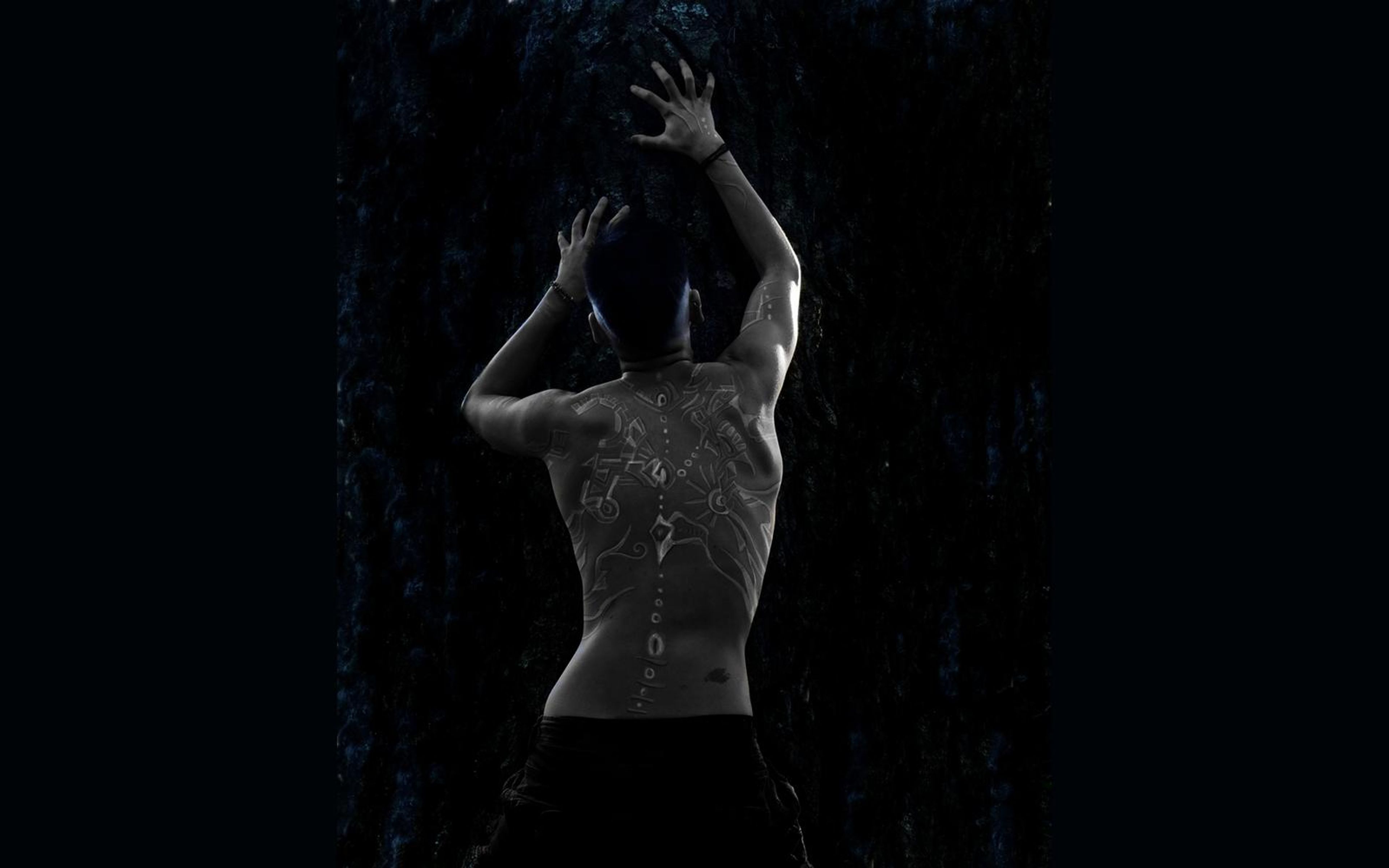 Man seen from behind with both hands raised against a dark textured wall, pale patterns painted on his back