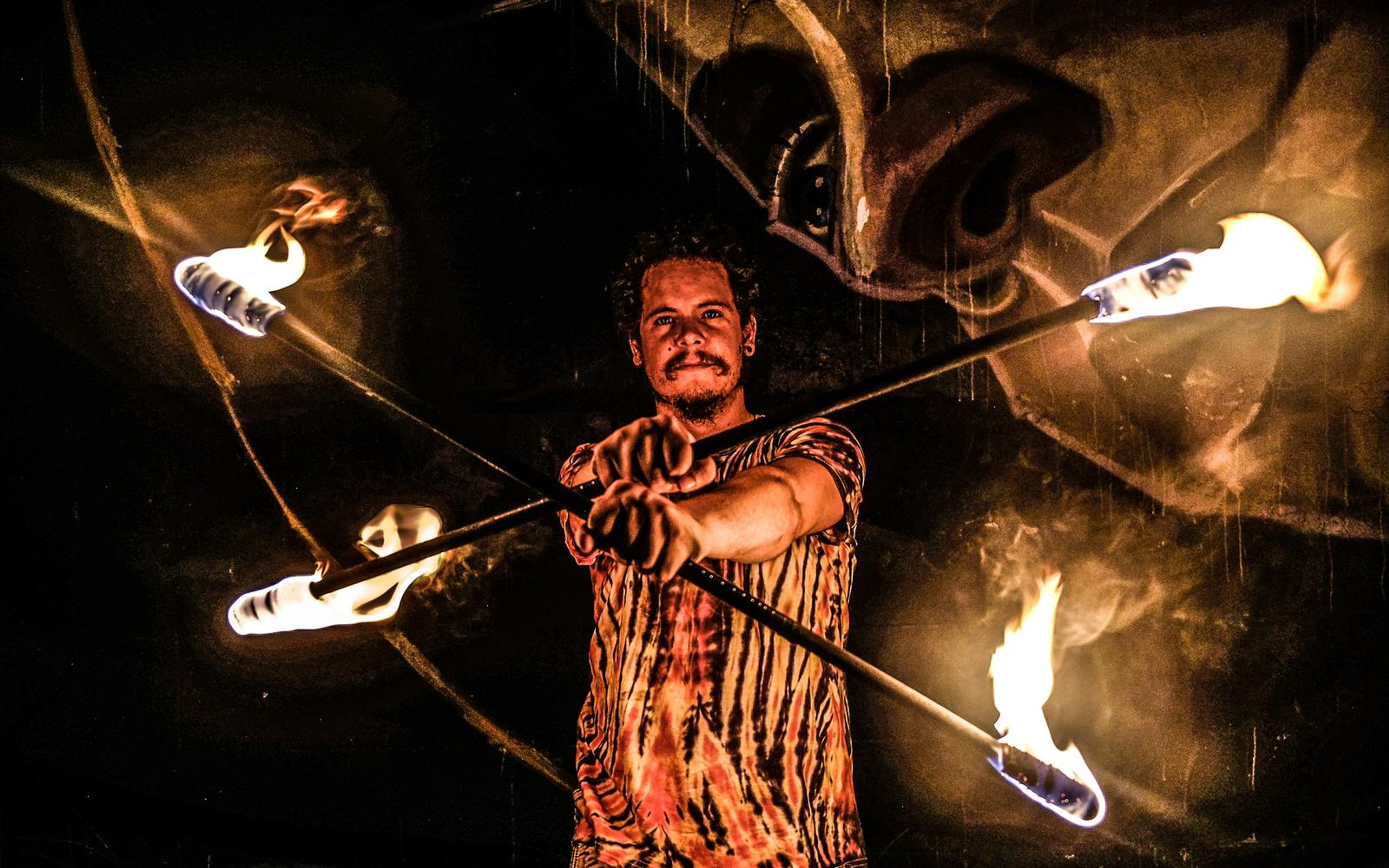Close view of a smiling man in a tie-dye shirt holding fire staffs, a painted face on the wall behind
