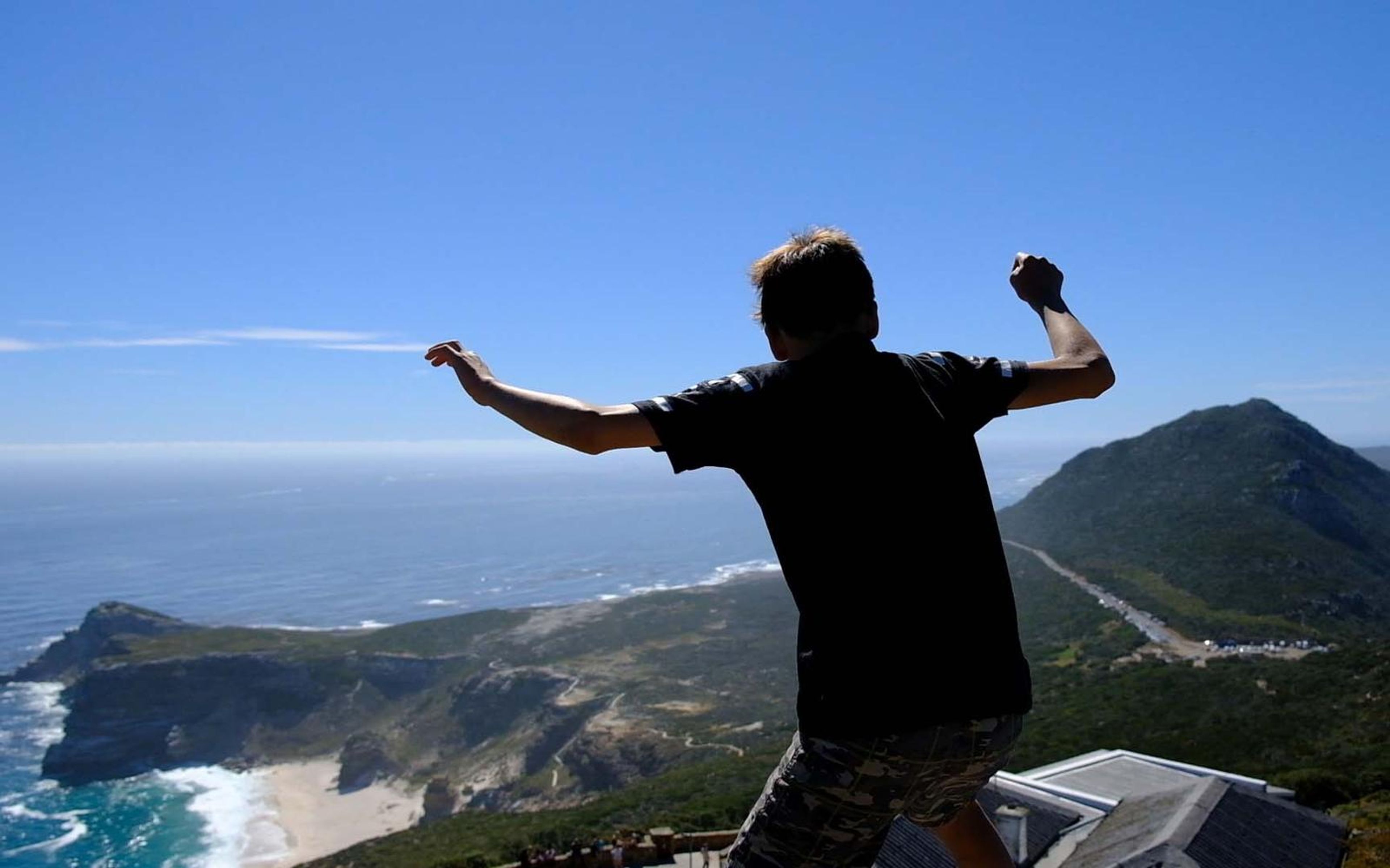 Boy in a dark shirt with arms thrown wide above a coastline of green headlands and sea