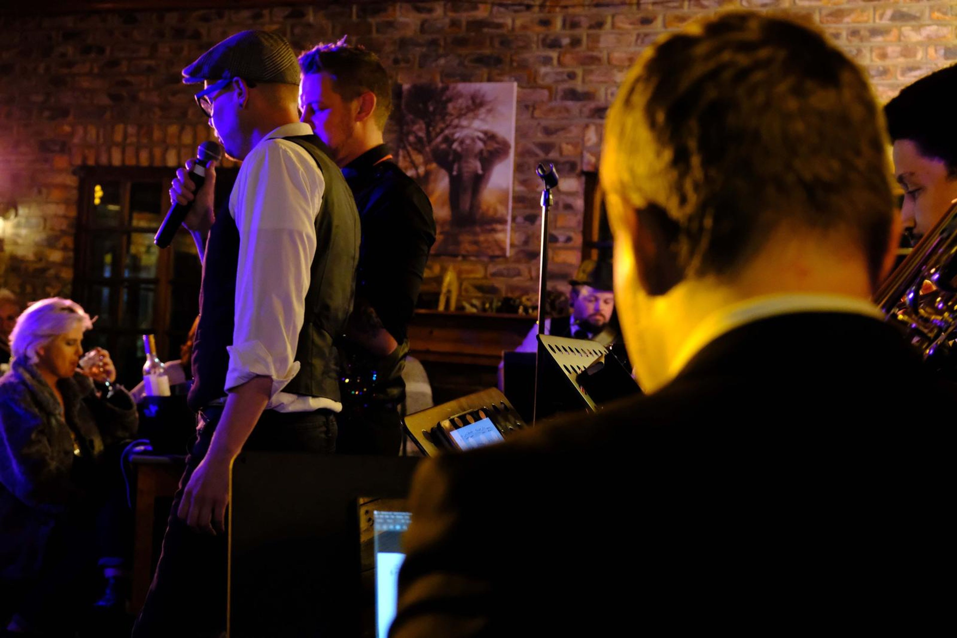 Singer in a waistcoat and flat cap standing beside the horn players
