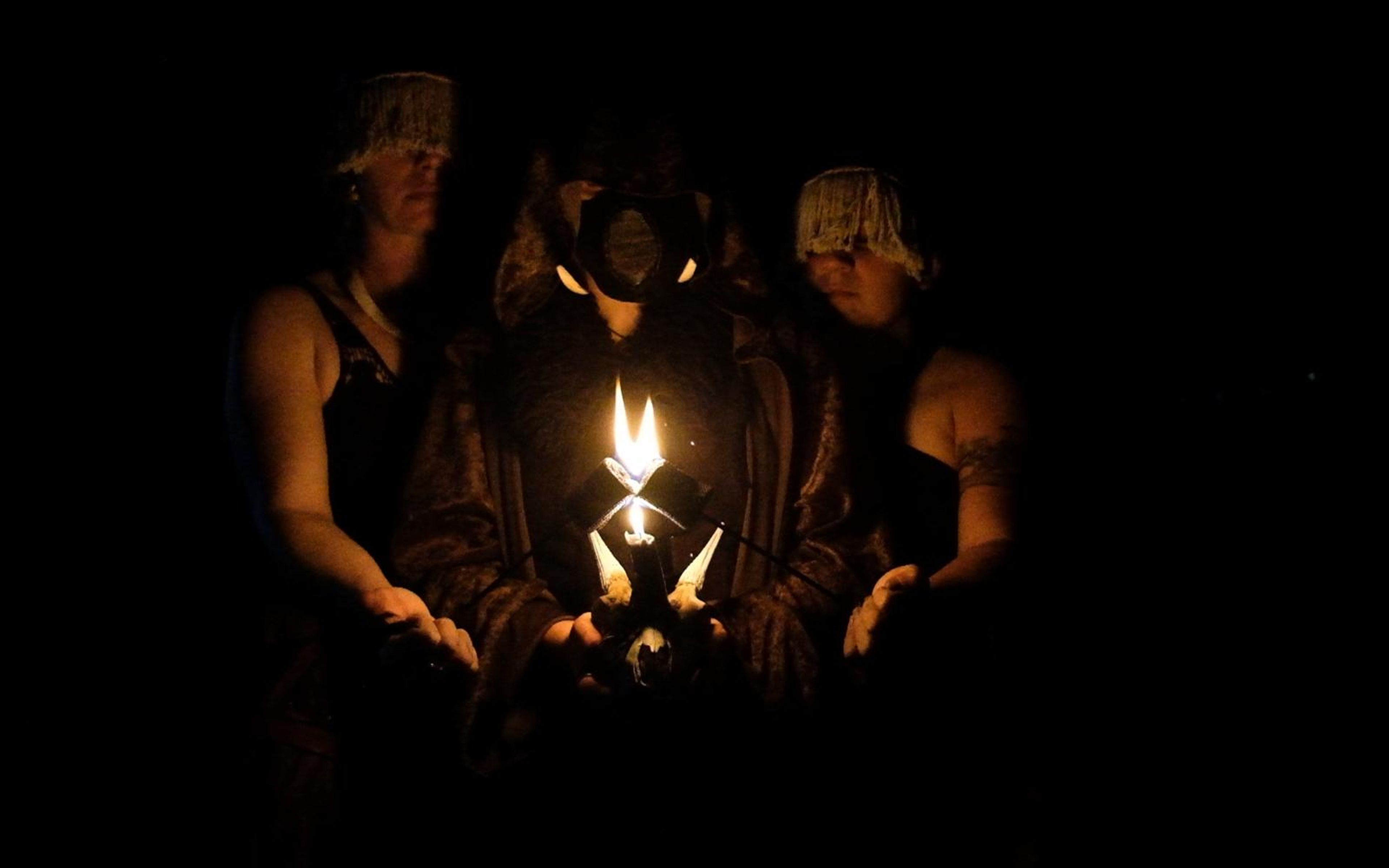 Three performers in a ritual line at night, small flame rising from a horned skull in the centre figure's hands