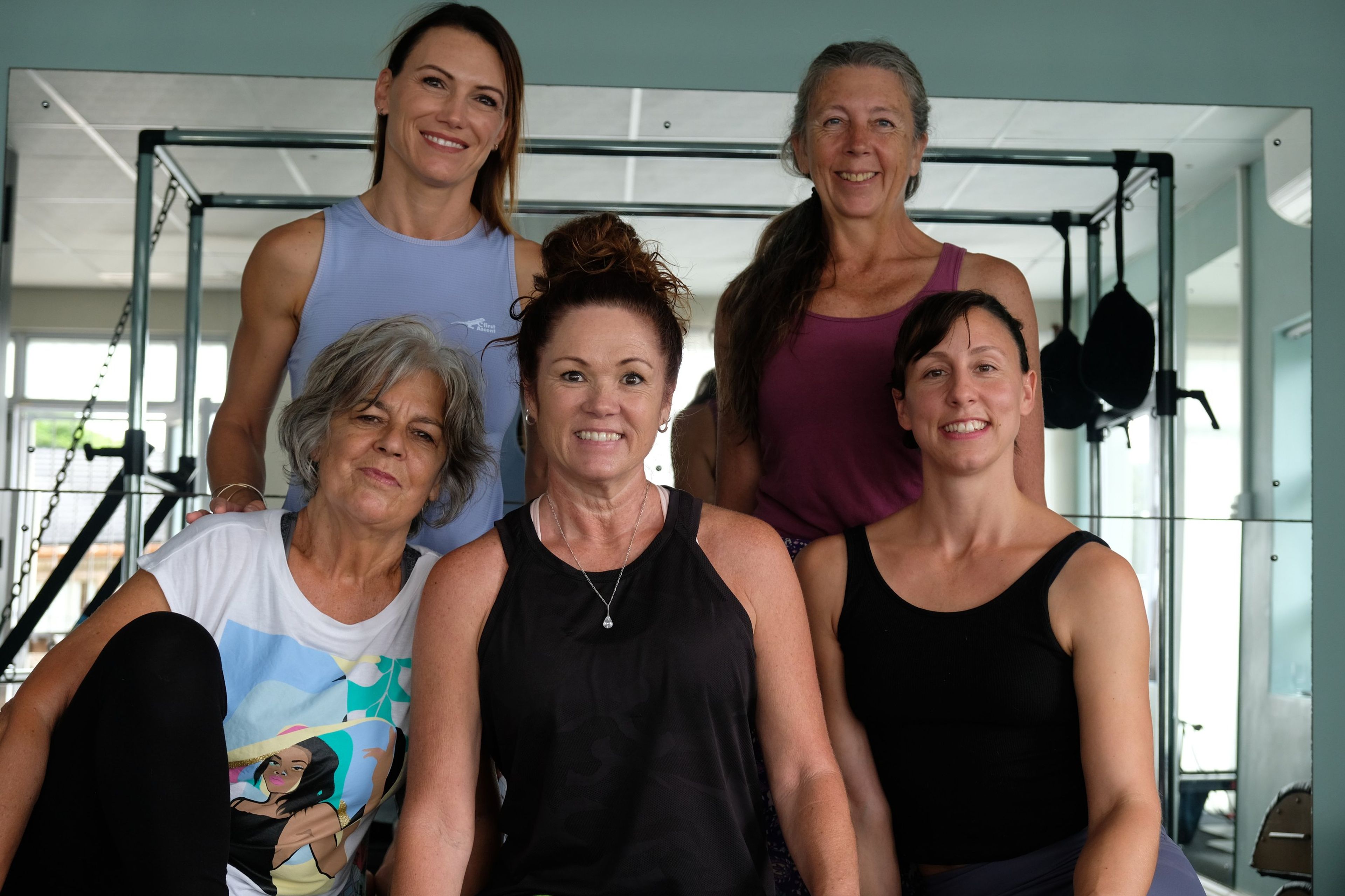 Five members of the Bend Pilates & Wellness Center team smiling together in two rows inside the studio, Pilates apparatus behind them