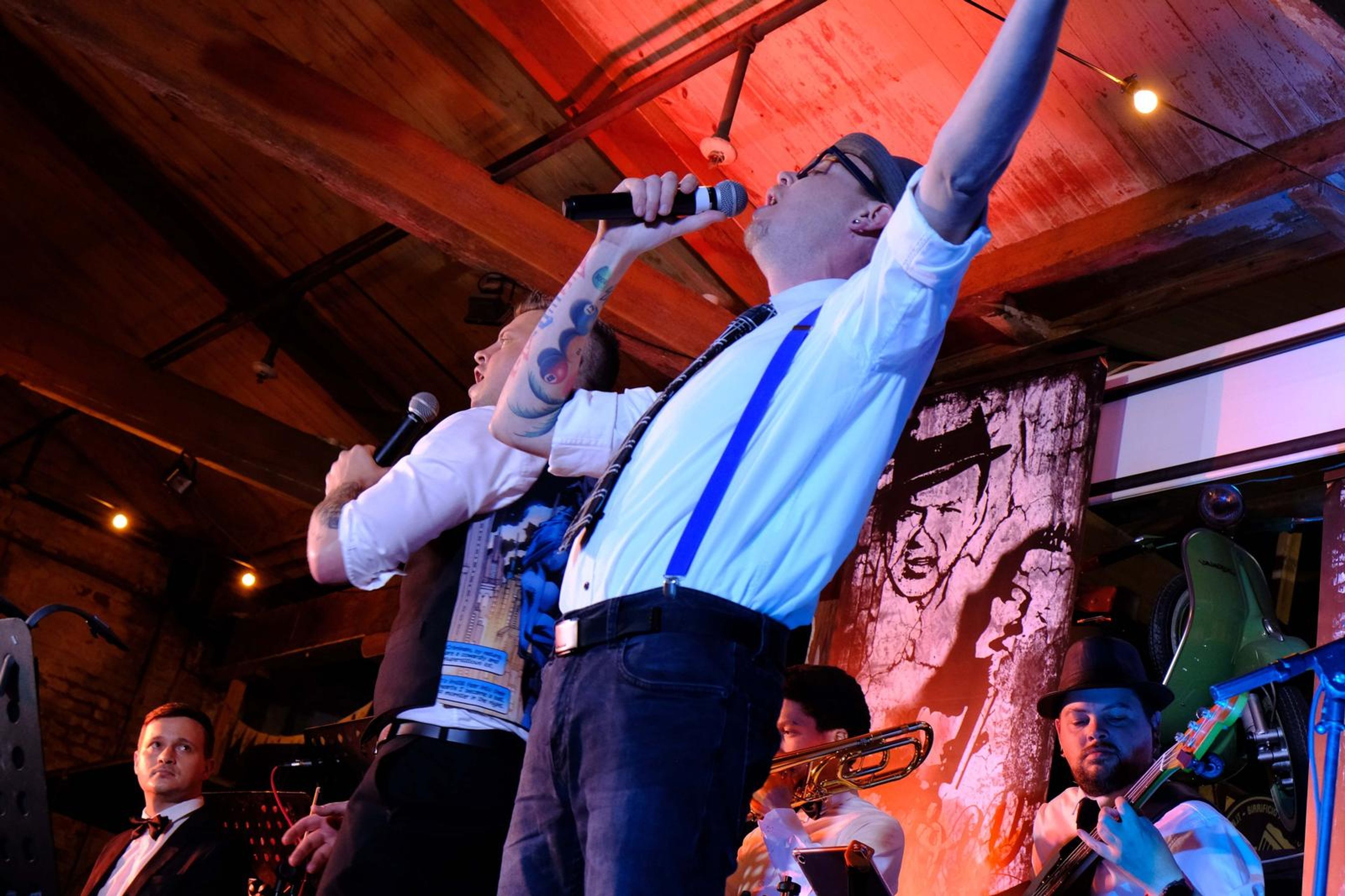 Singer in a hat and suspenders reaching up with the microphone