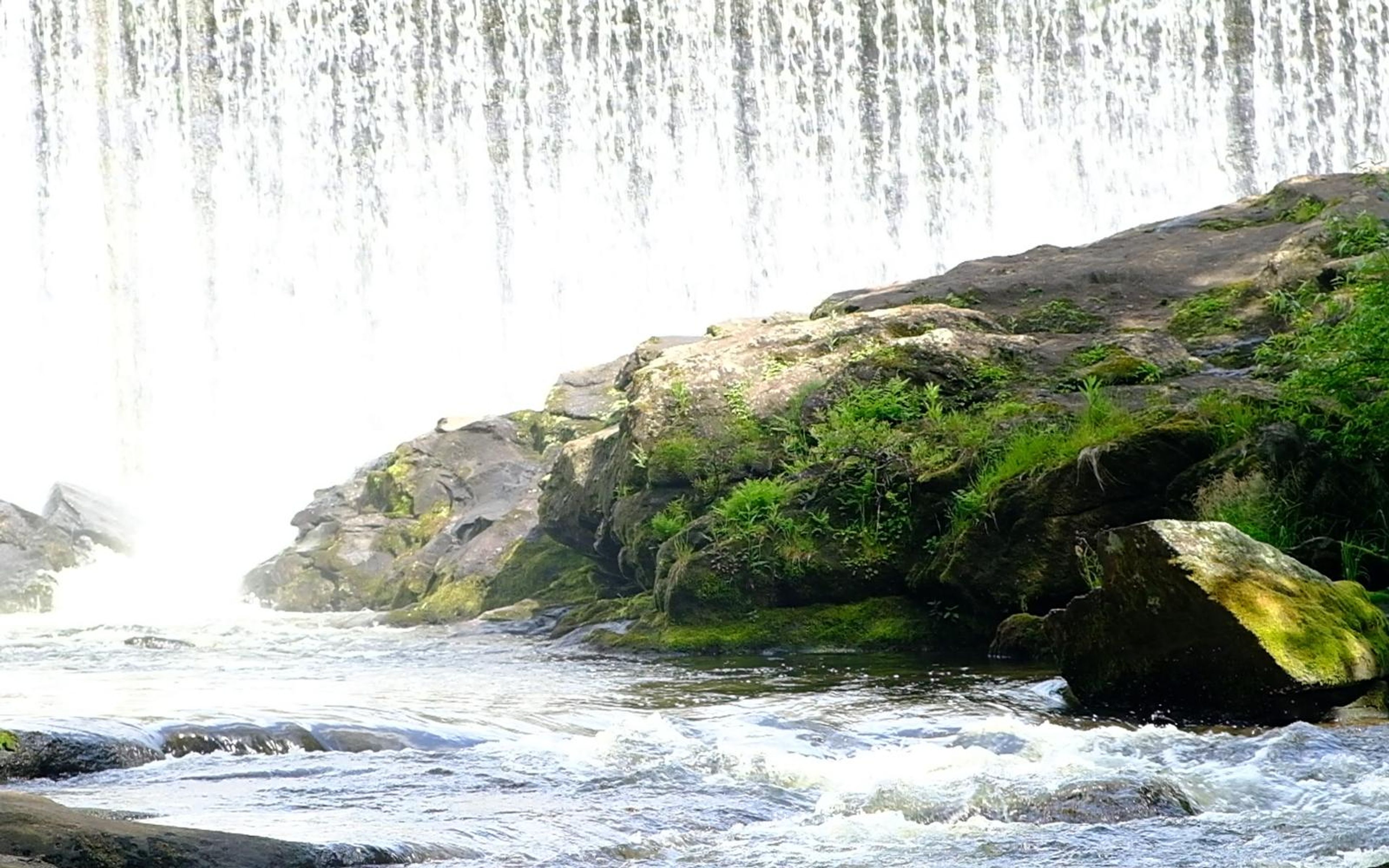 Bright water curtain spilling beside a mossy boulder and rapids in full sun