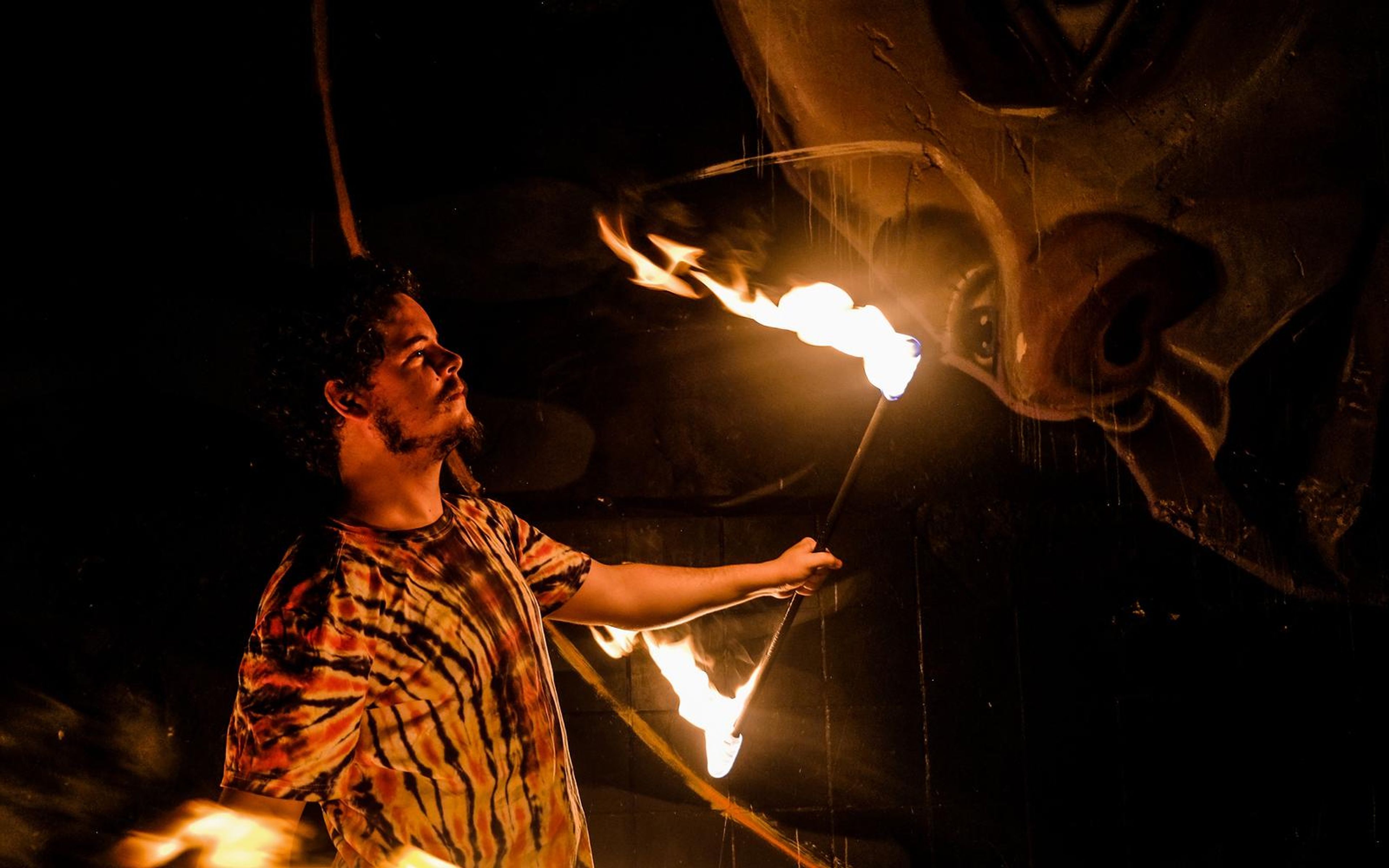 Man in profile holding a burning staff out to the side, large painted faces on the wall behind