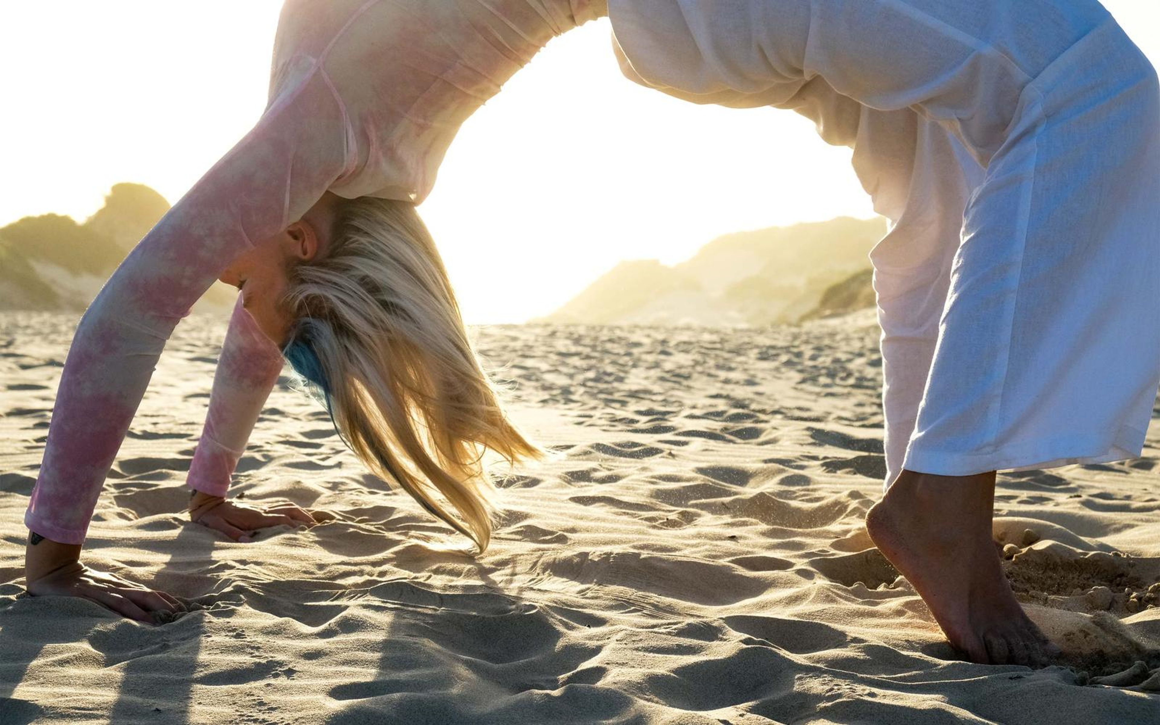 Woman in a backbend arch over the sand with her hair brushing the ground and sunlit dunes behind