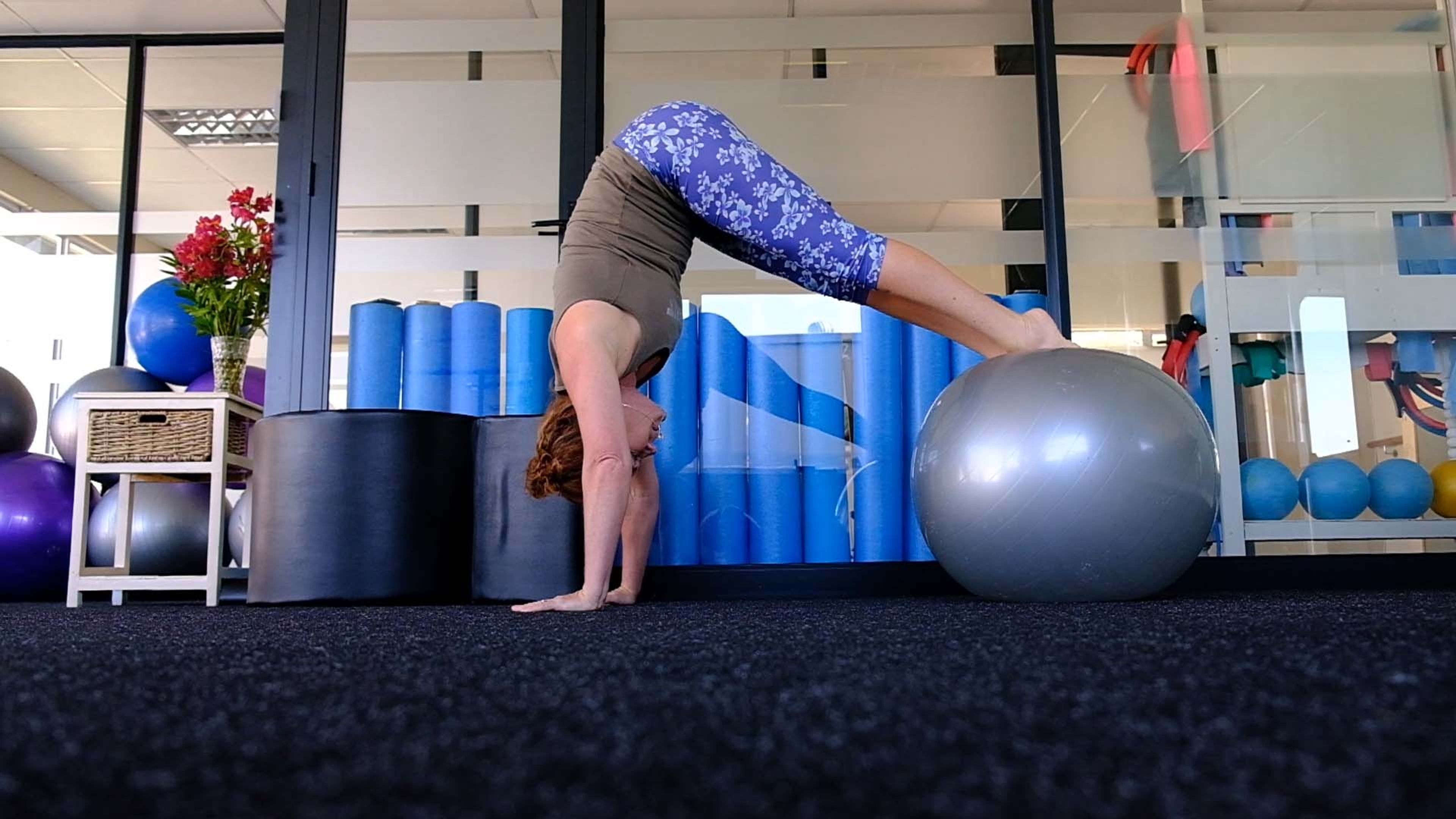 A woman in a handstand-style pose over a grey stability ball in front of foam rollers