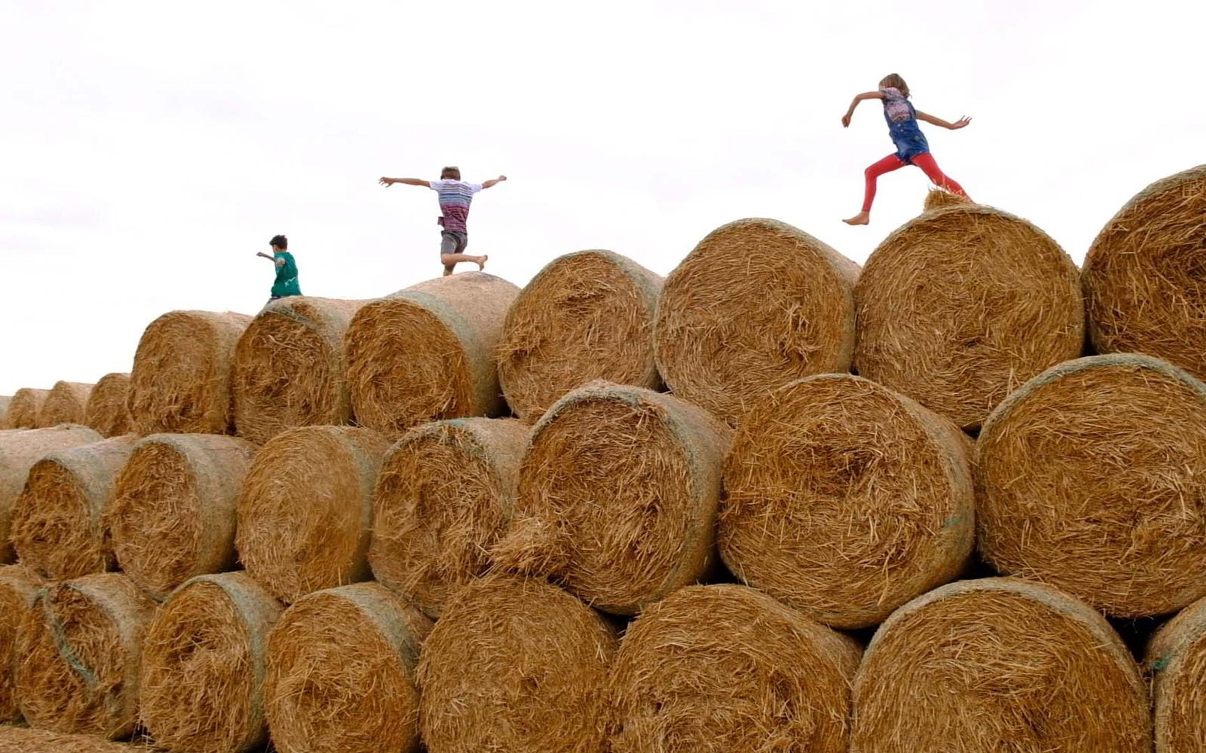Two children leaping across a stack of round hay bales against a bright white sky