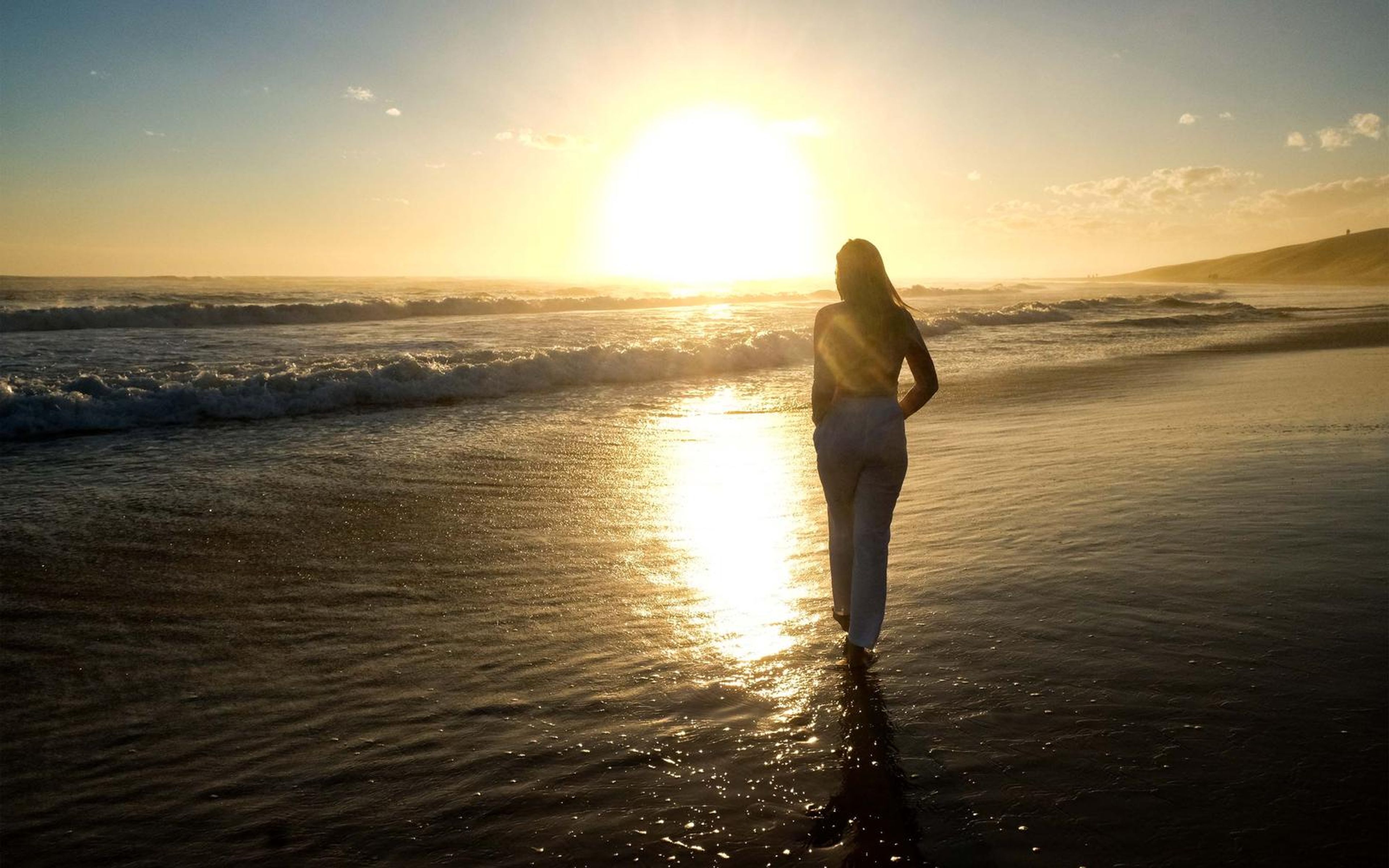 Silhouette of a woman walking into the sea toward a low setting sun, wet sand reflecting gold light