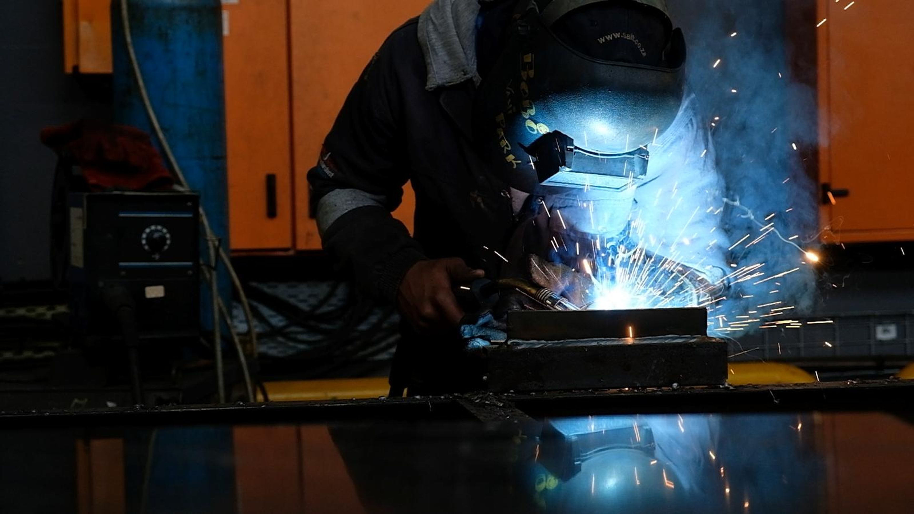 Welder in a mask bent over a steel bar with smoke and sparks rising