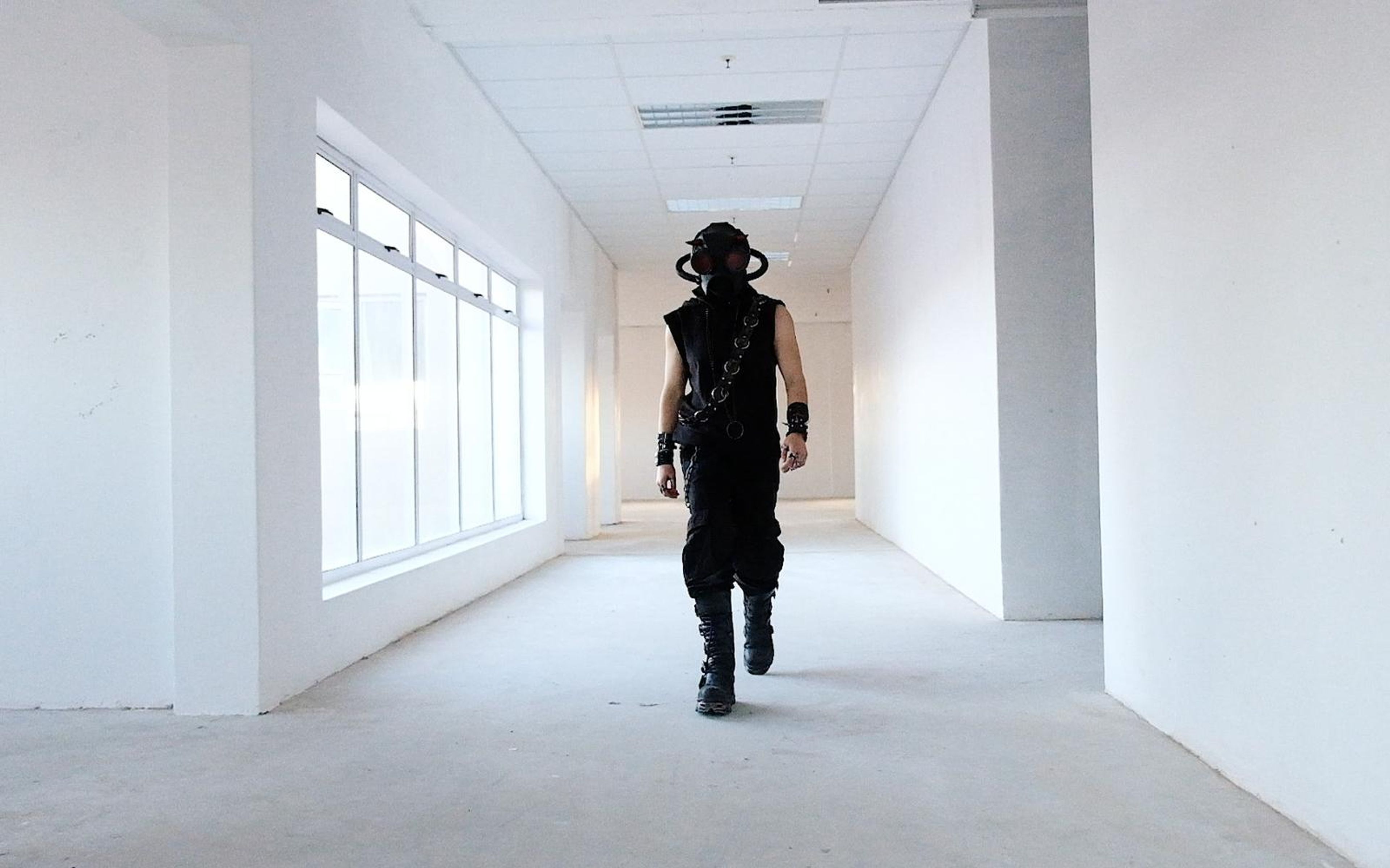 Figure in a black horned gas mask with red lenses and chained black outfit standing near tall windows in a white hallway