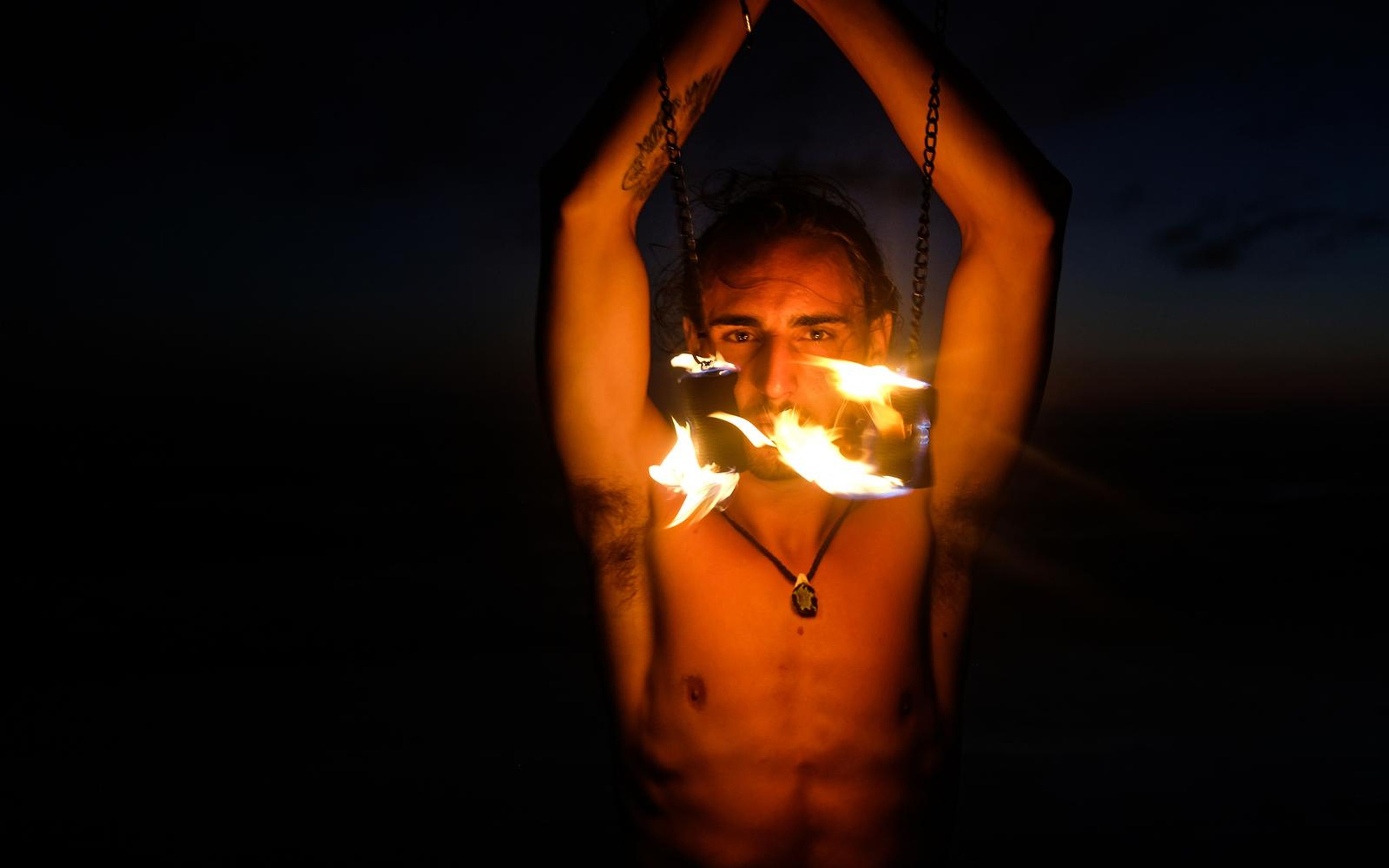 Man framed by his raised arms with flames at both shoulders and warm orange light on his face