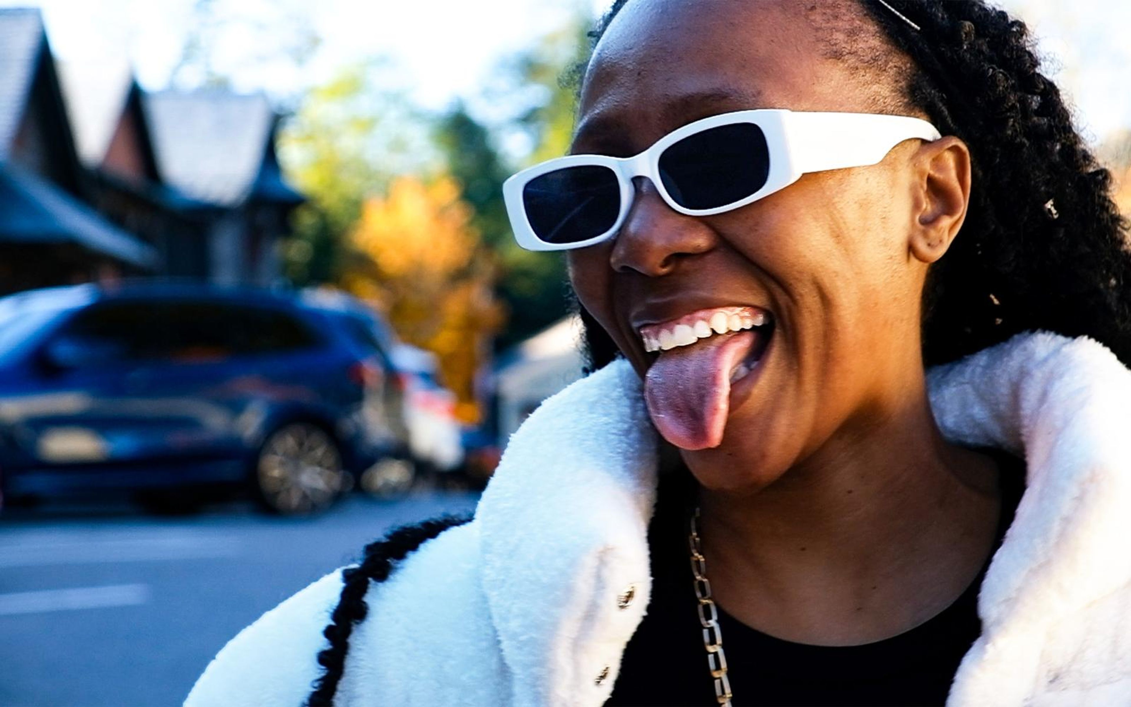 Model sticking out her tongue playfully, white rectangular sunglasses, fur jacket, blurred street behind