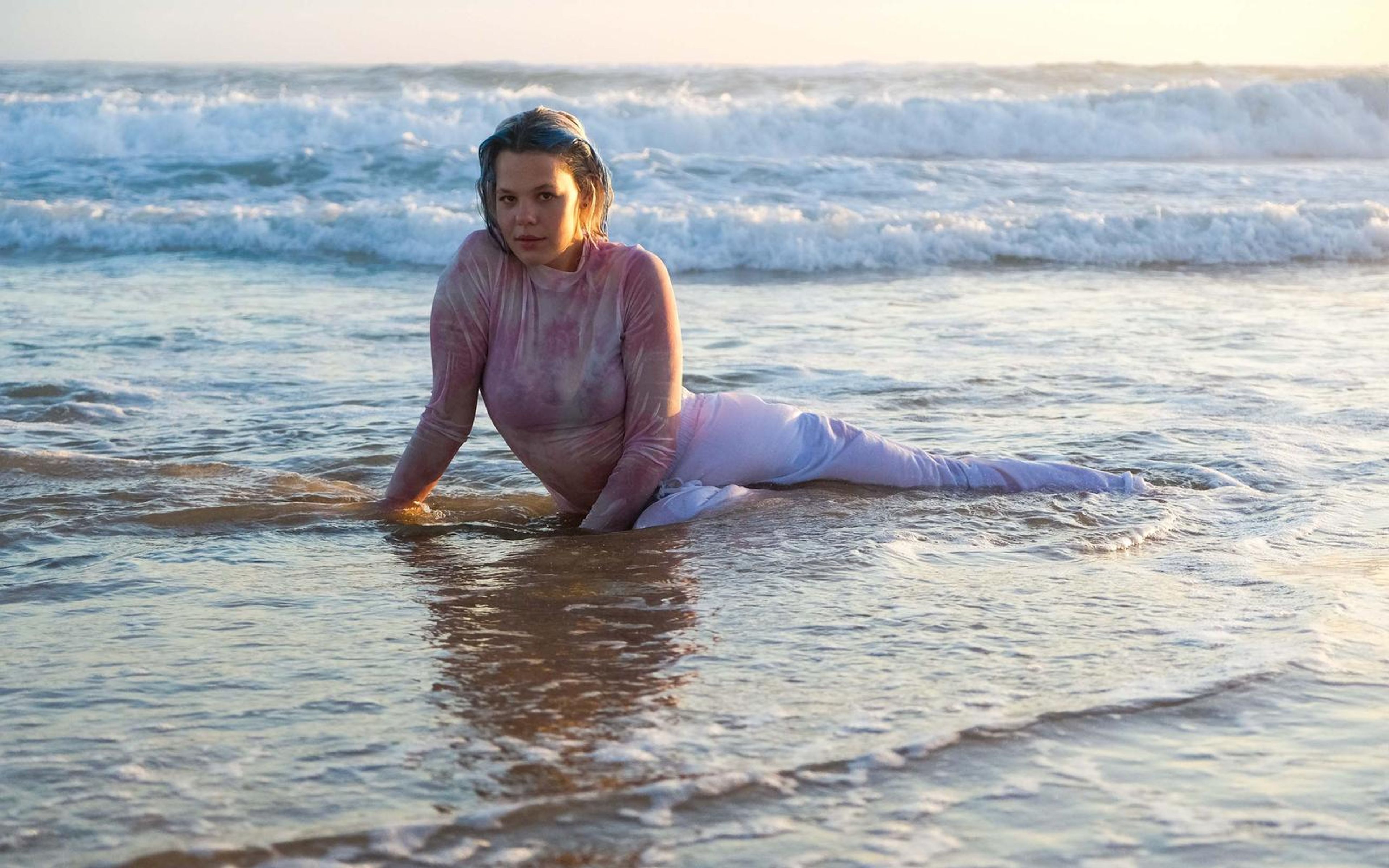 Woman propped on her arm in thin tide water at dusk, white trousers trailing, glowing reflection beneath her