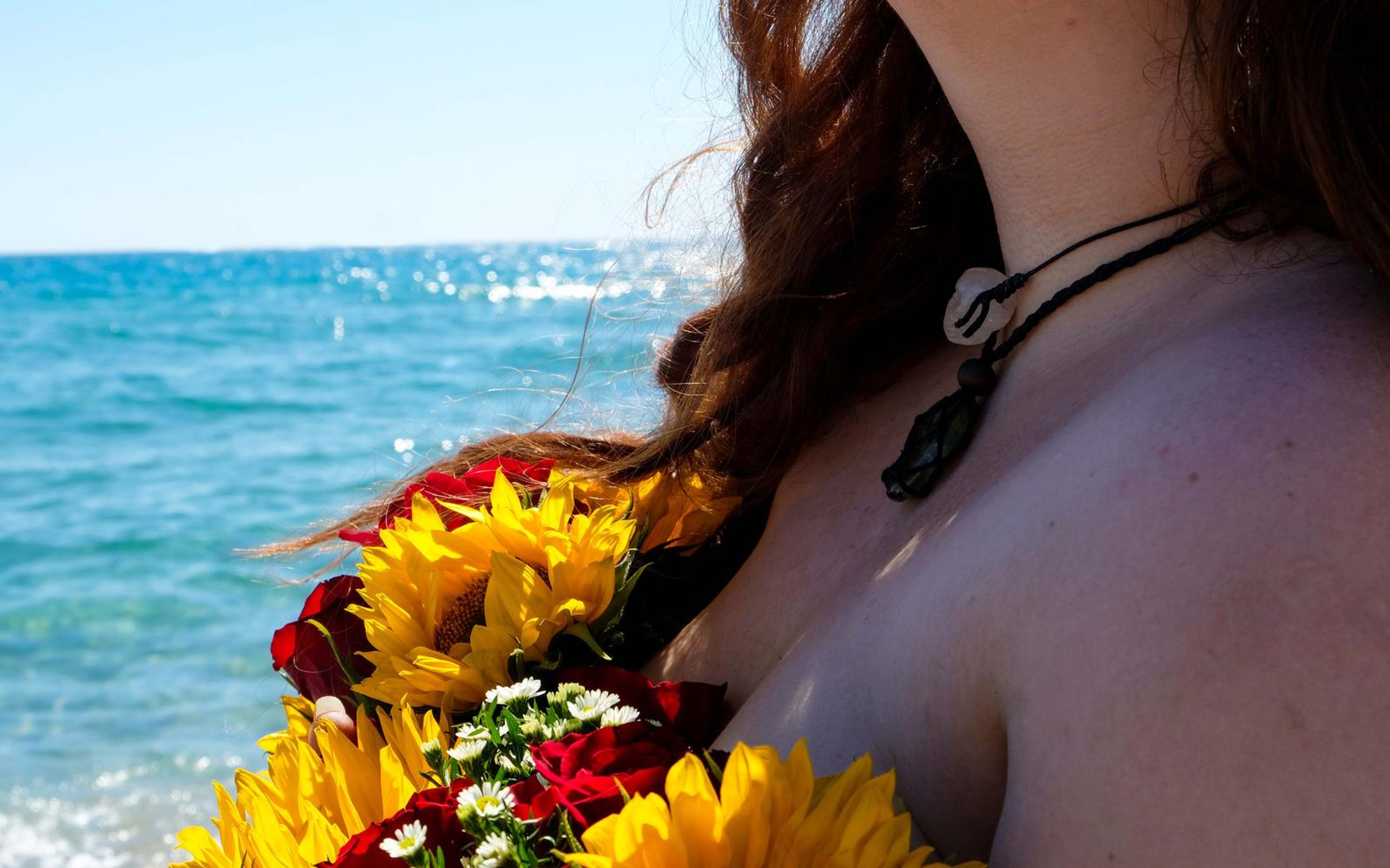Tight crop of sunflowers and red roses held at the collarbone, a braided cord pendant necklace, soft horizon and sunlit sea in the background