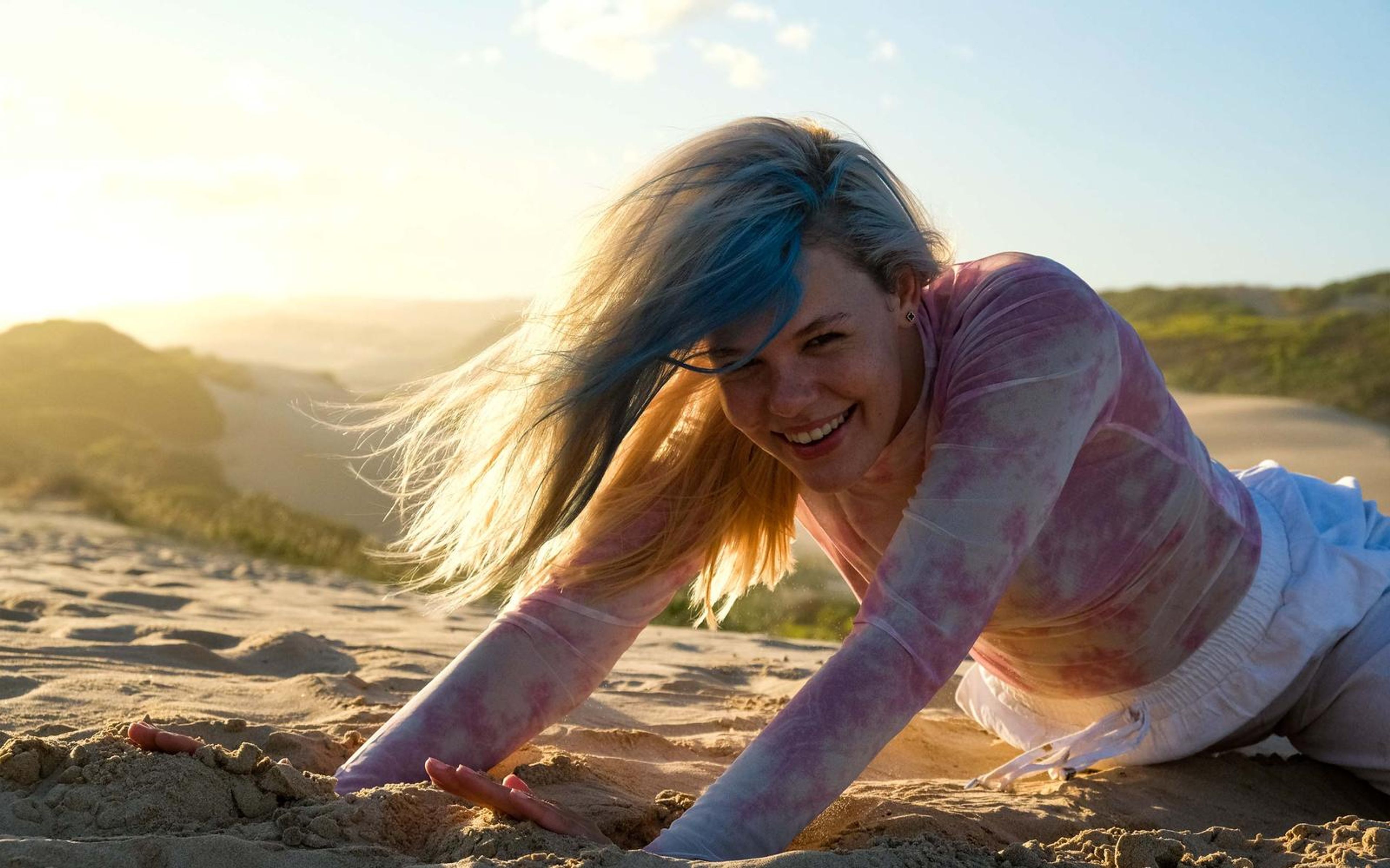 Woman smiling and leaning forward on her hands in the sand with wind in her hair