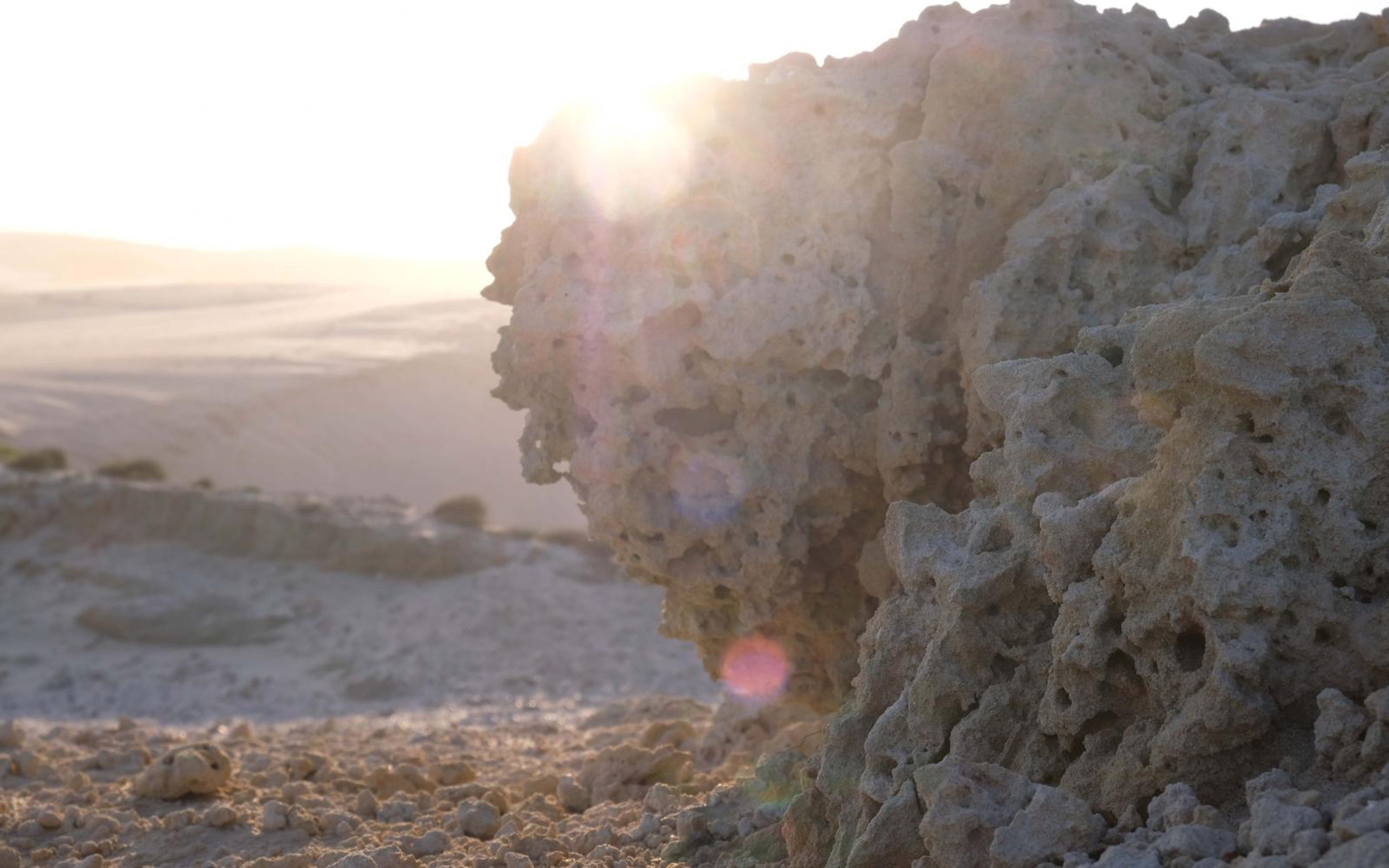 Sun flaring over the edge of a textured limestone outcrop above the dunes
