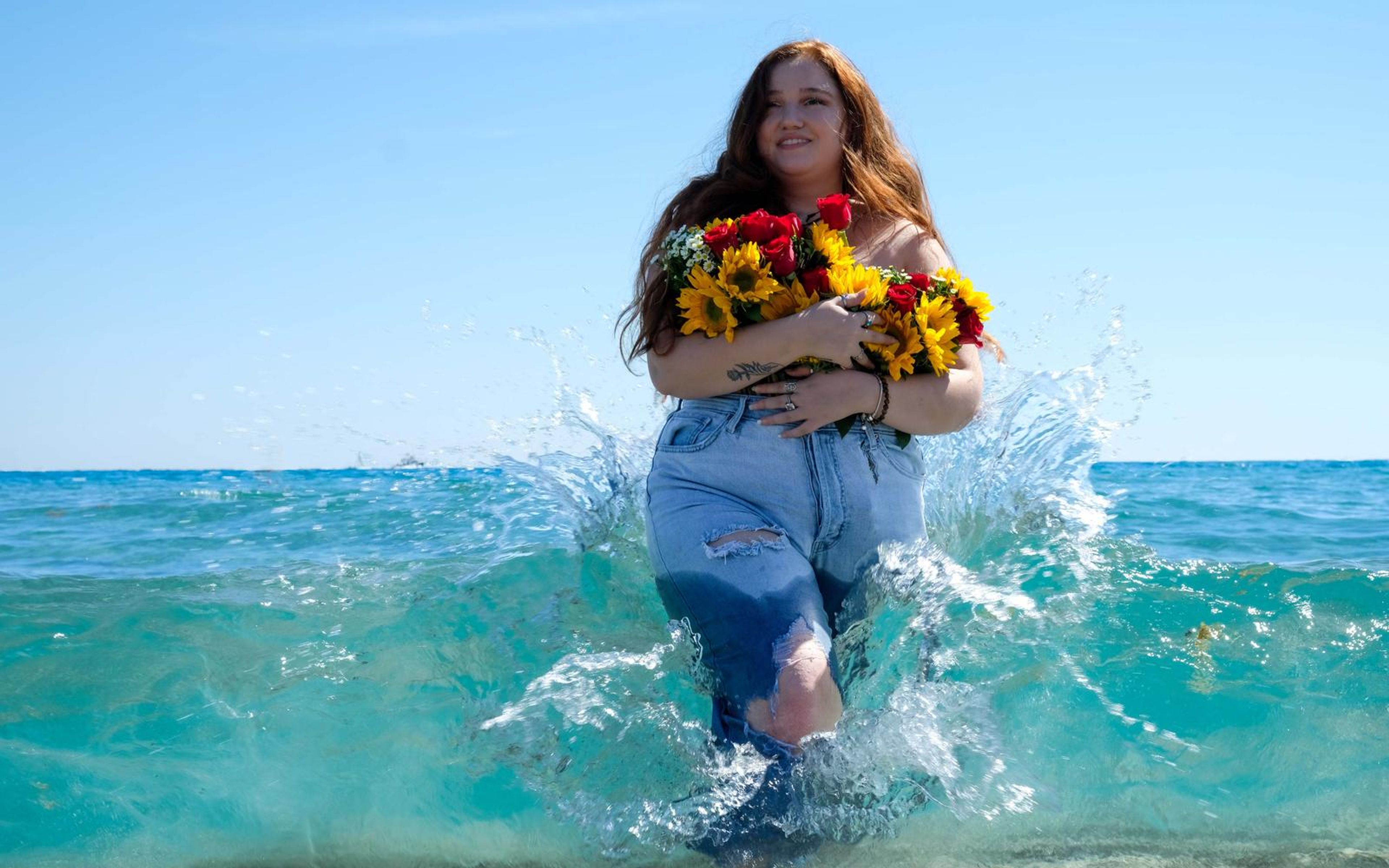 Model walking toward camera through splashing water, smiling, bouquet in her arms, wet denim