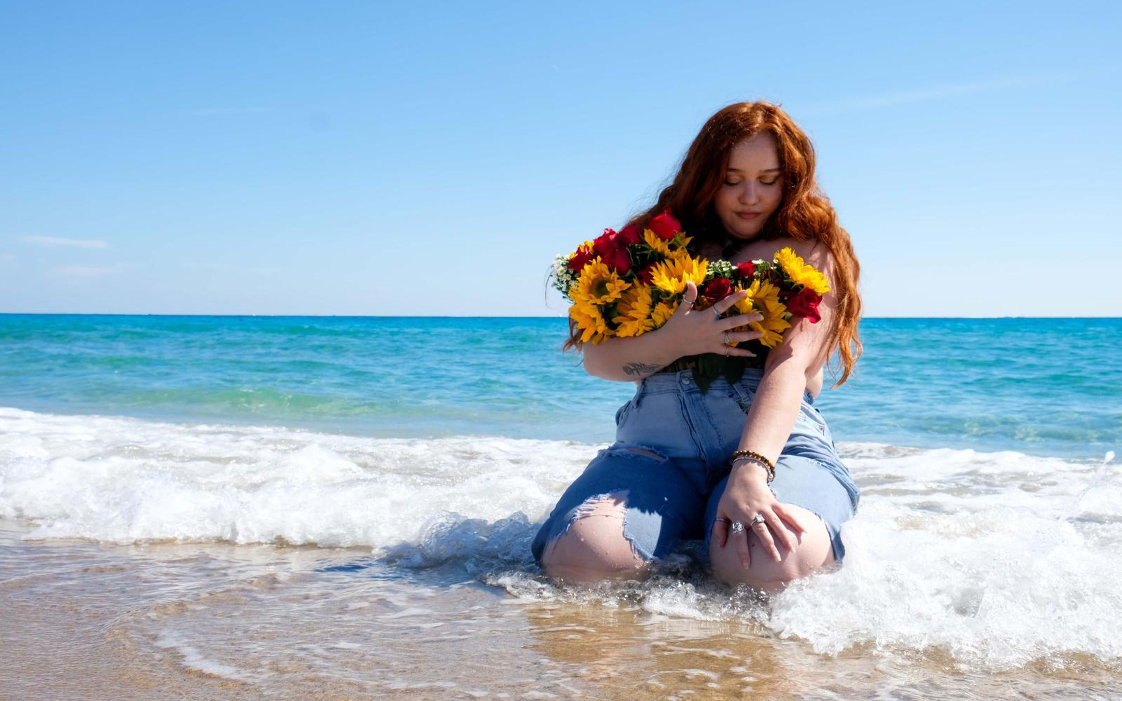 Model kneeling in shallow surf holding sunflowers and red roses, denim overalls, bright blue sky behind