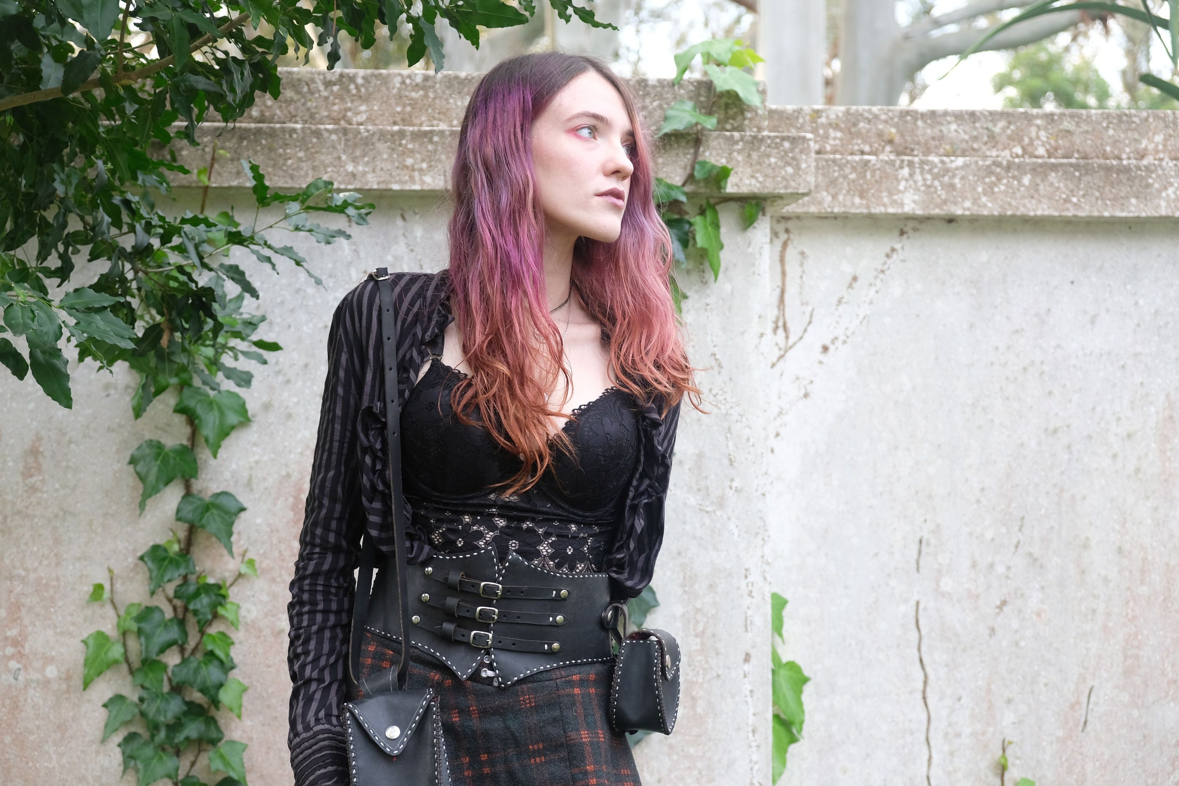 Woman with pink hair in a black corset belt and plaid skirt standing by an ivy wall