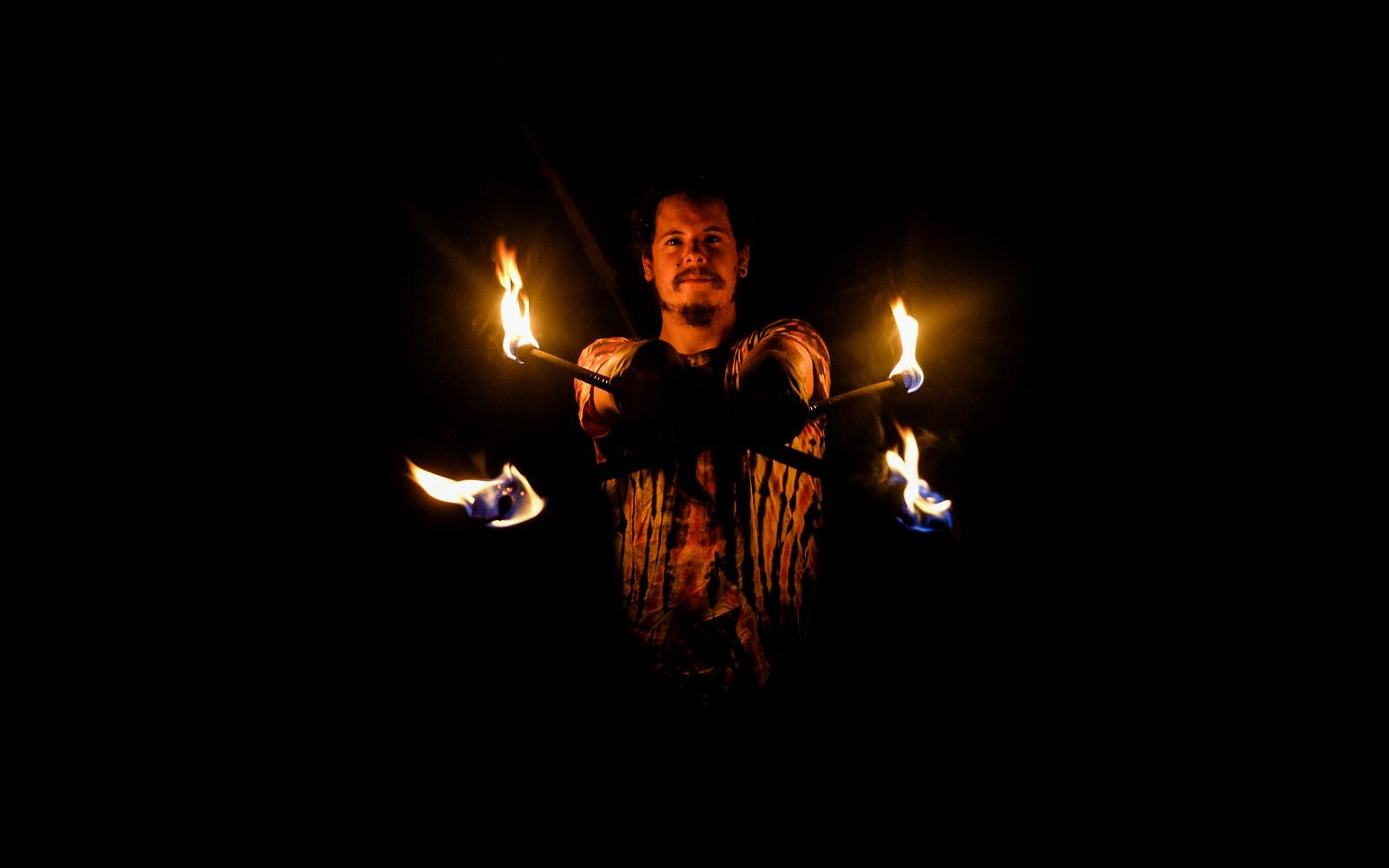 Distant view of a man in a patterned shirt surrounded by four small flames in the dark