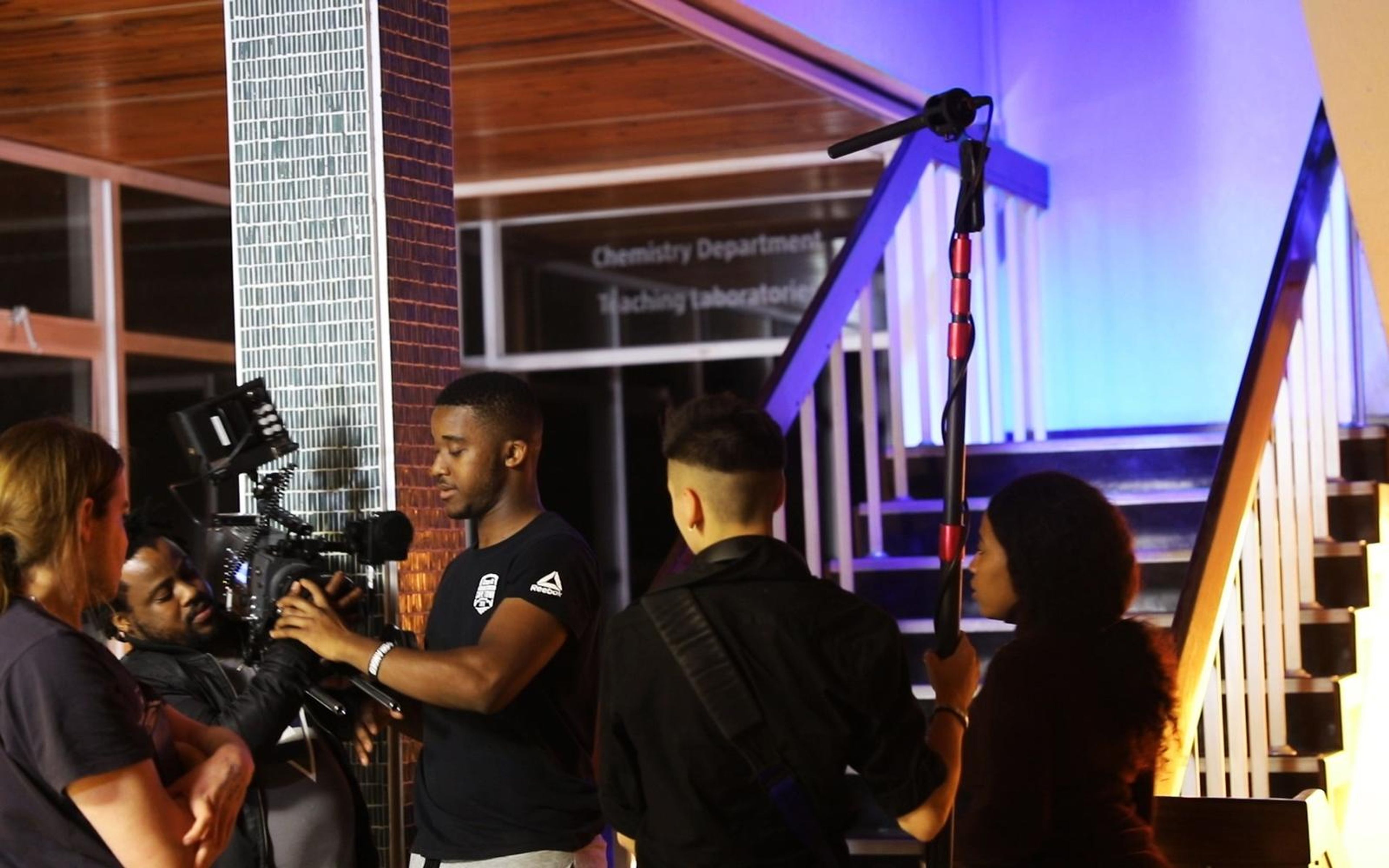 Camera operator and boom operator filming in a lit stairwell, with crew gathered beside them