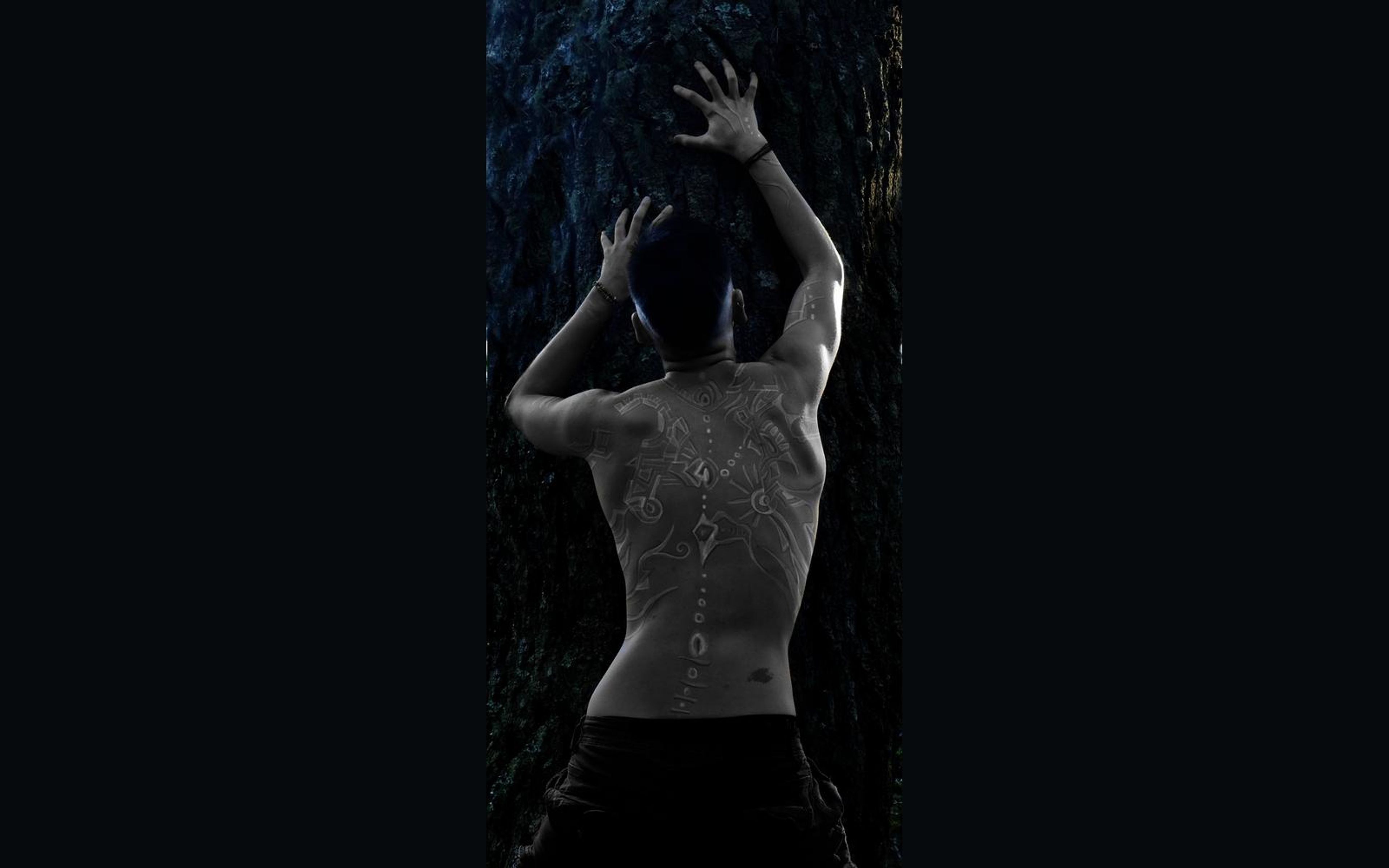 Tall narrow frame of a figure from behind with raised hands, painted glyphs along the spine, deep shadow