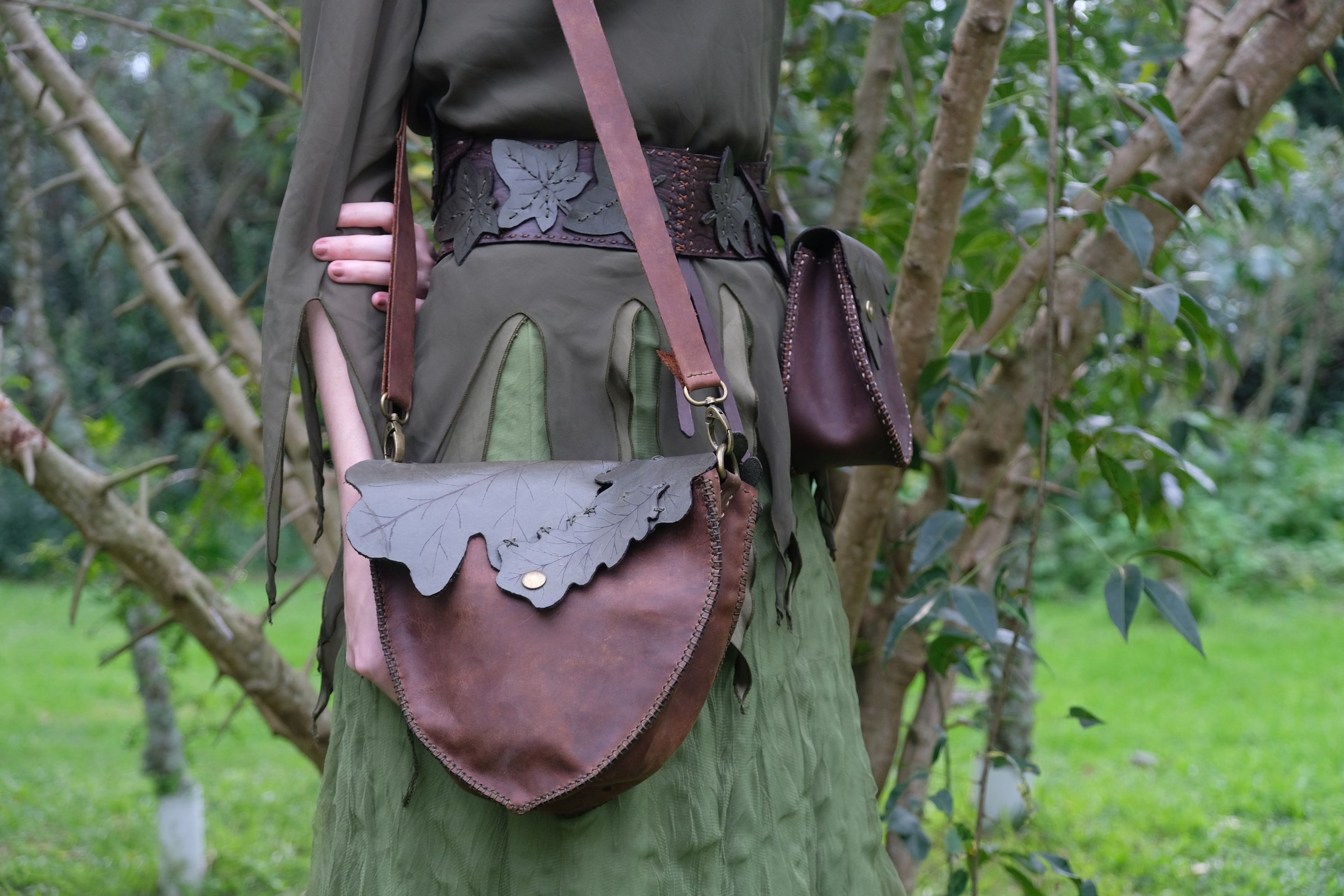Brown leather bag with a grey leaf-shaped flap, in front of green trees