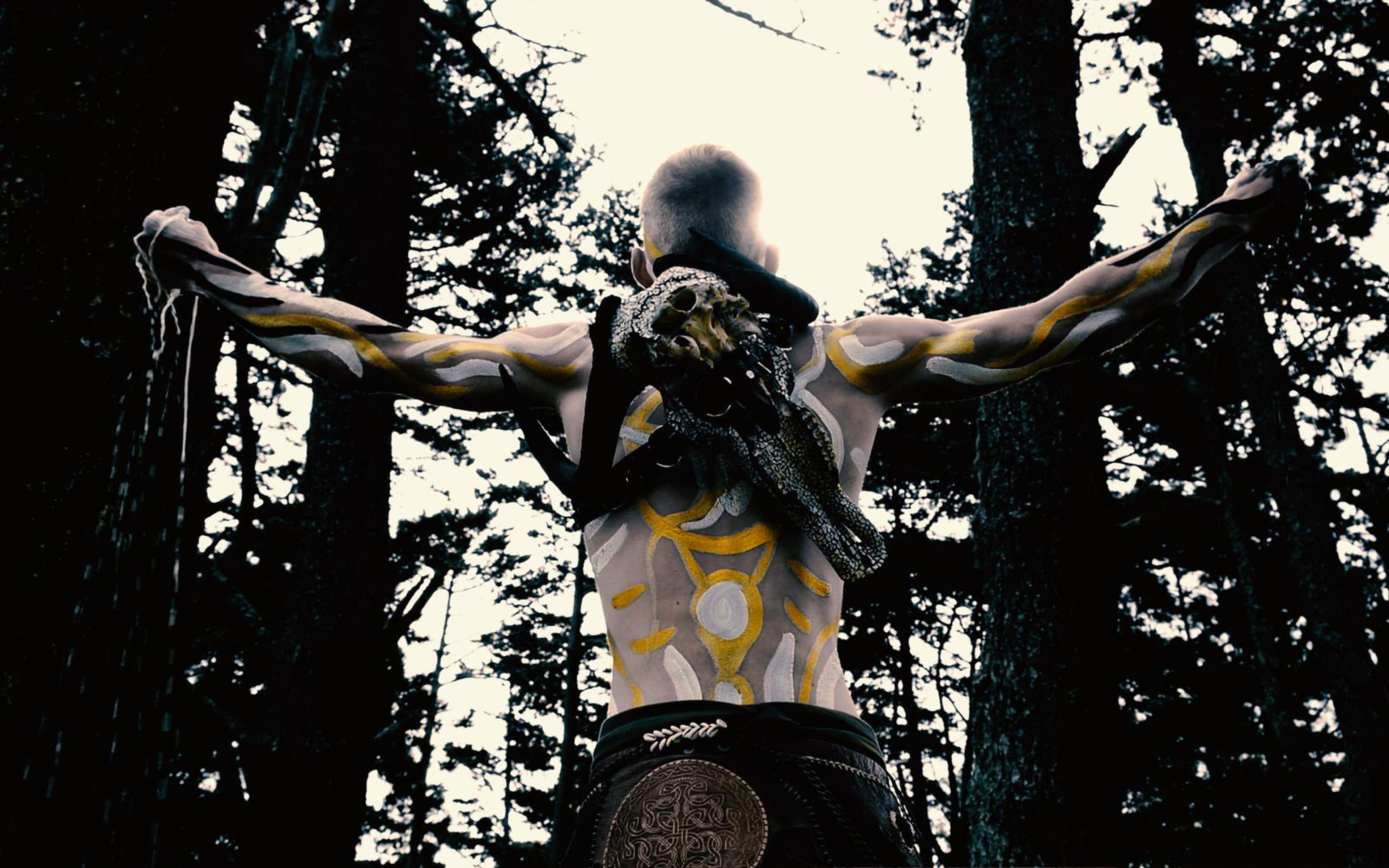 Back view of a body-painted performer with arms spread in a pine forest, a bull skull strapped to the back, pale sky behind