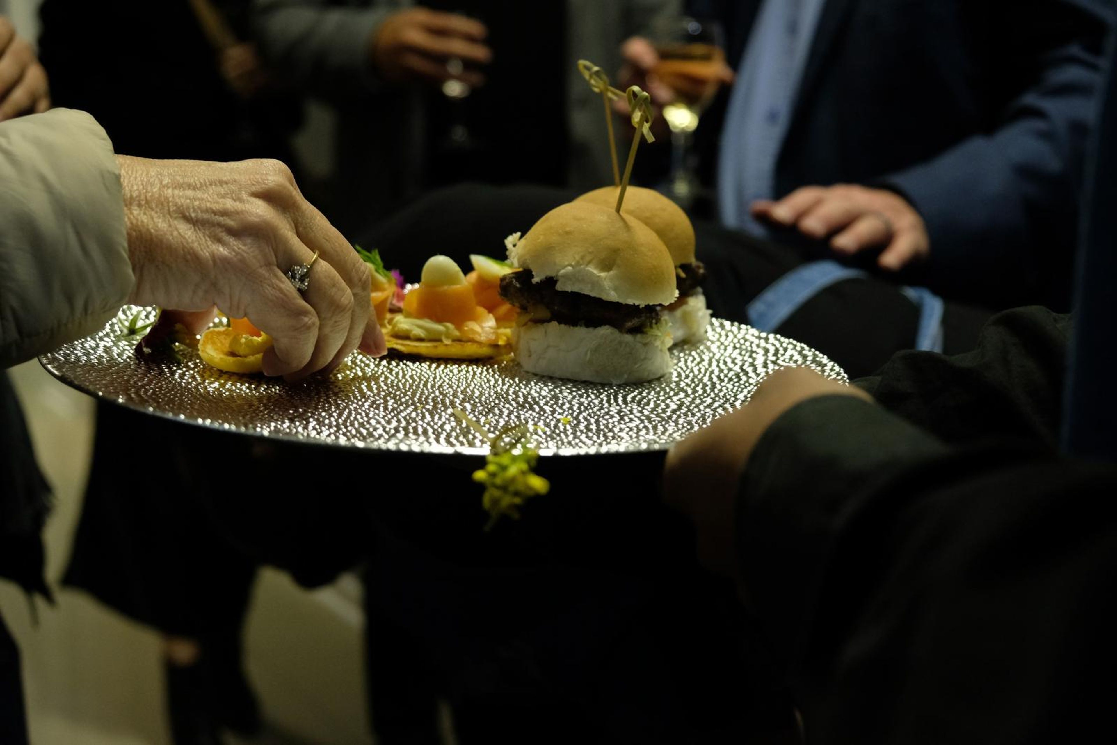 Hand taking a slider from a platter of mini burgers and smoked salmon blinis
