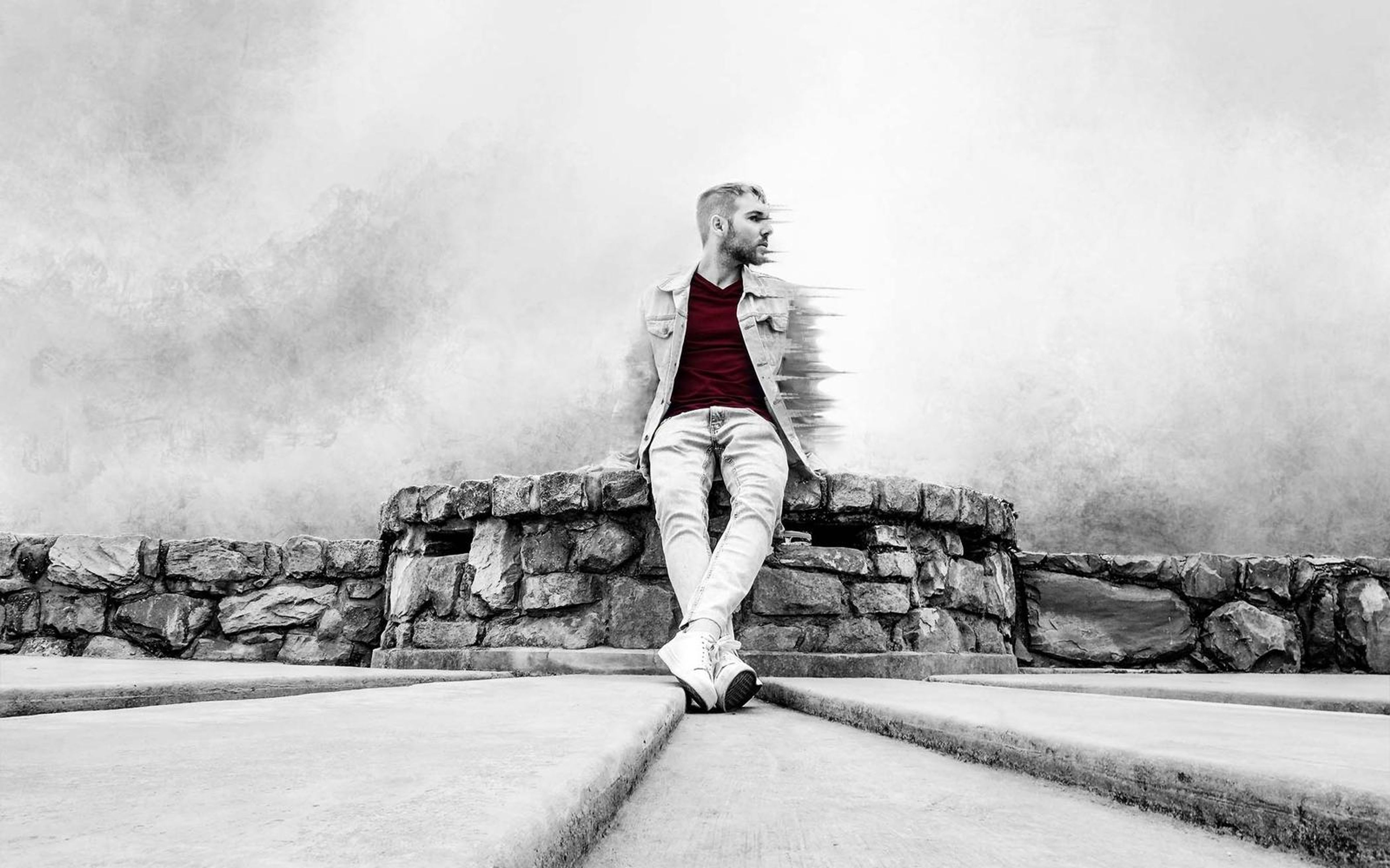 Man in a pale denim jacket and maroon tee seated on a stone wall, his edge dissolving into grey sea spray with colour kept only in his shirt