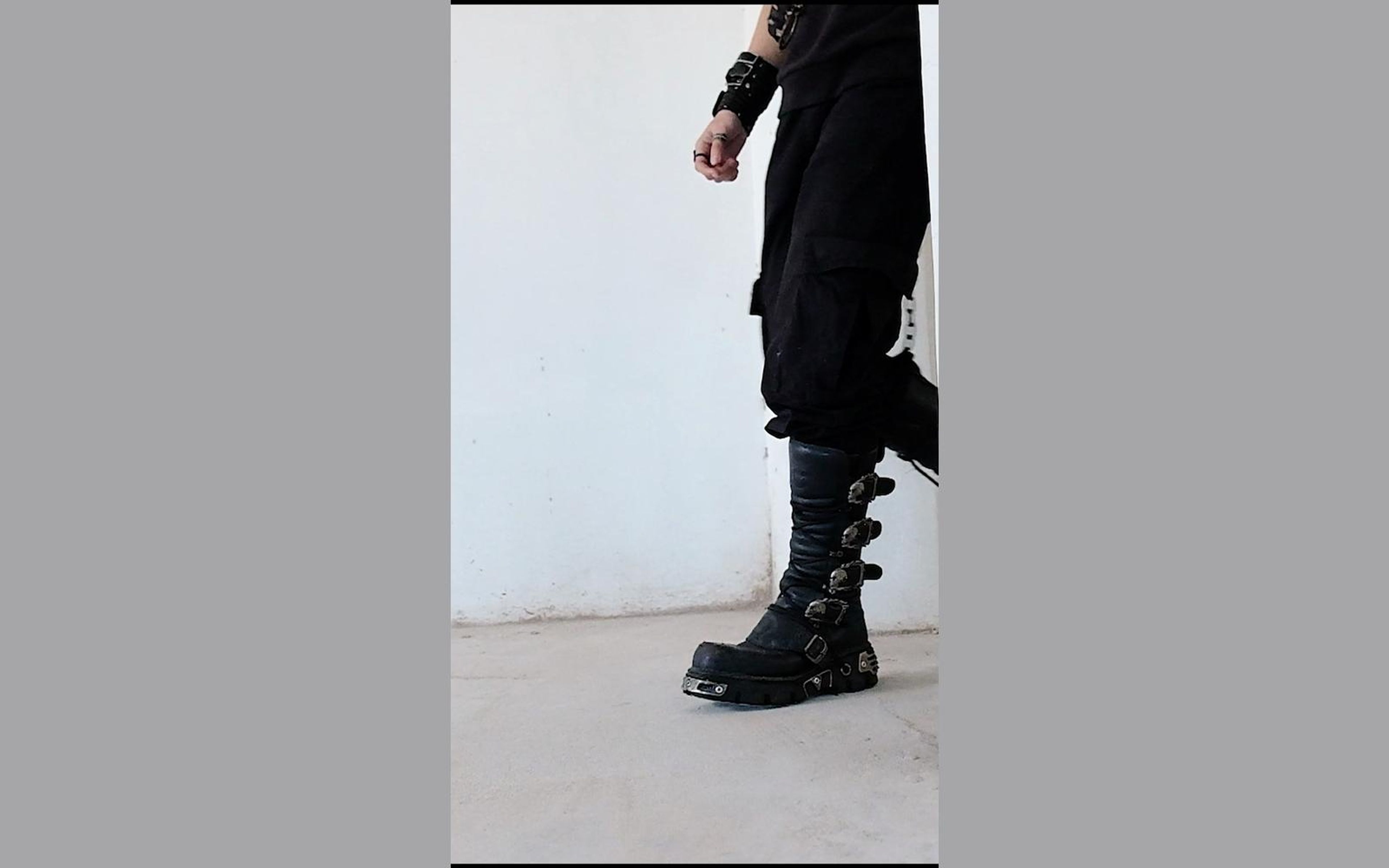 Clenched hand at the figure's side above a buckled New Rock boot against a white wall