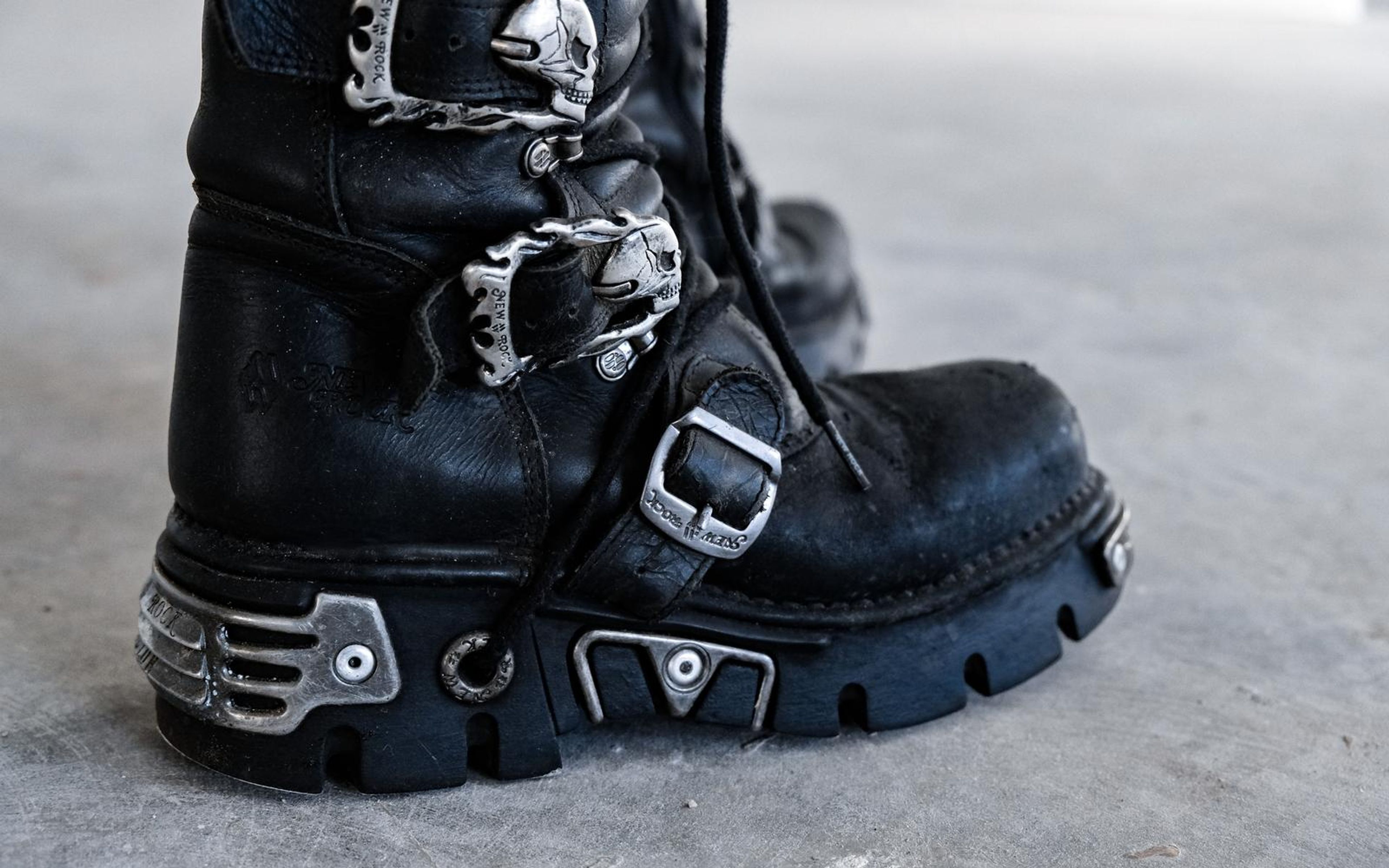 Close view of a black boot heel with skull buckles and metal plates on grey concrete, soft focus background