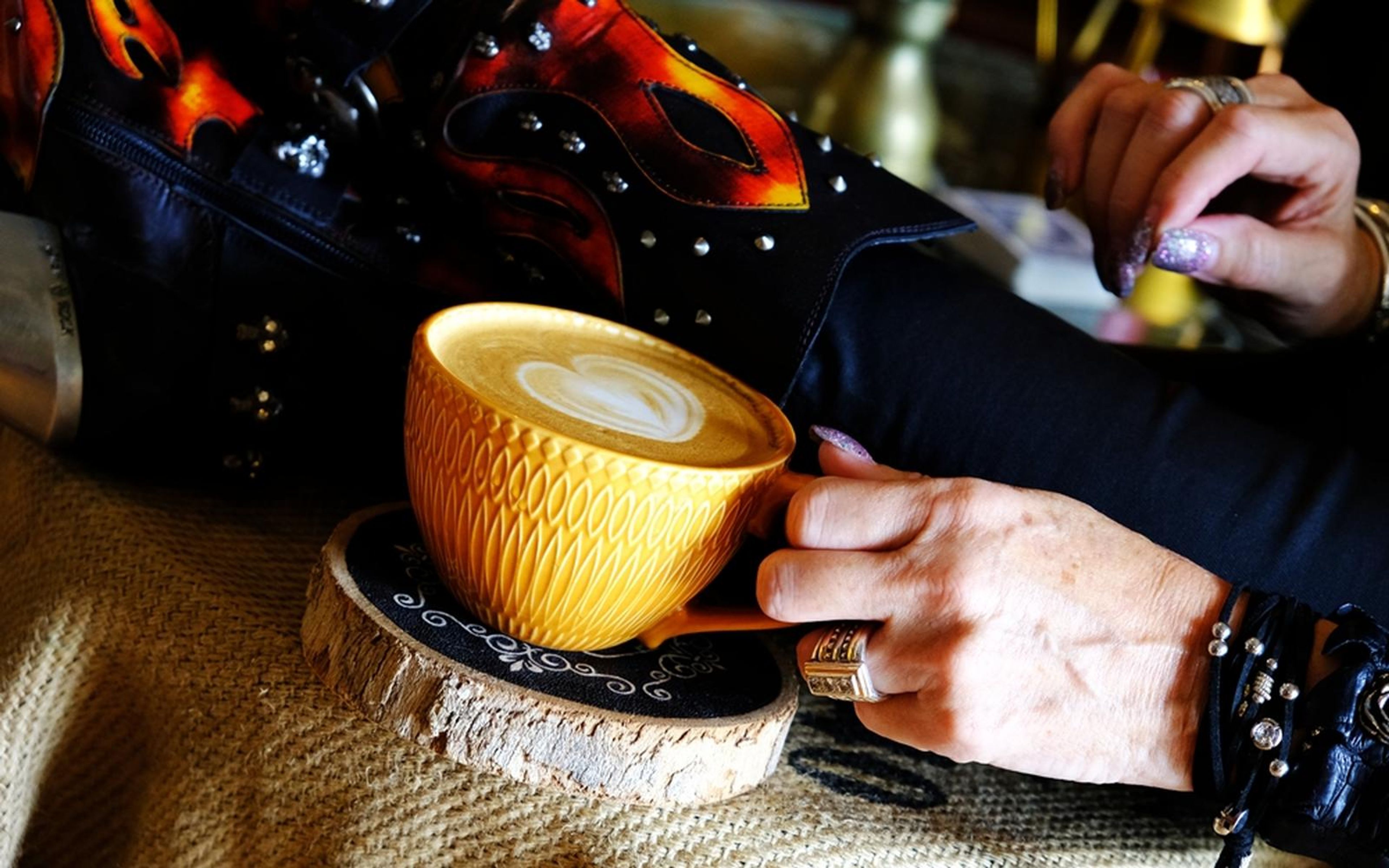 Golden cup of heart-shaped latte art resting beside a flame-patterned boot, hand on knee