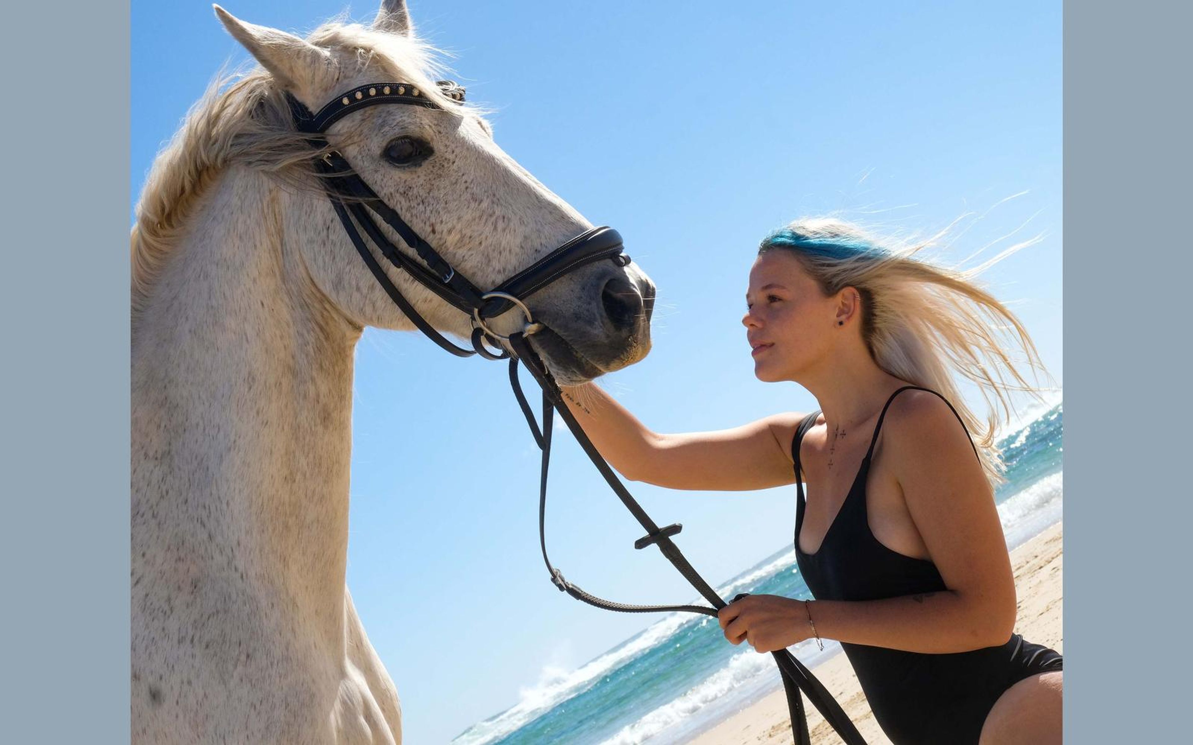 Woman in a black swimsuit holding the reins and looking at a white horse against bright blue sky and surf