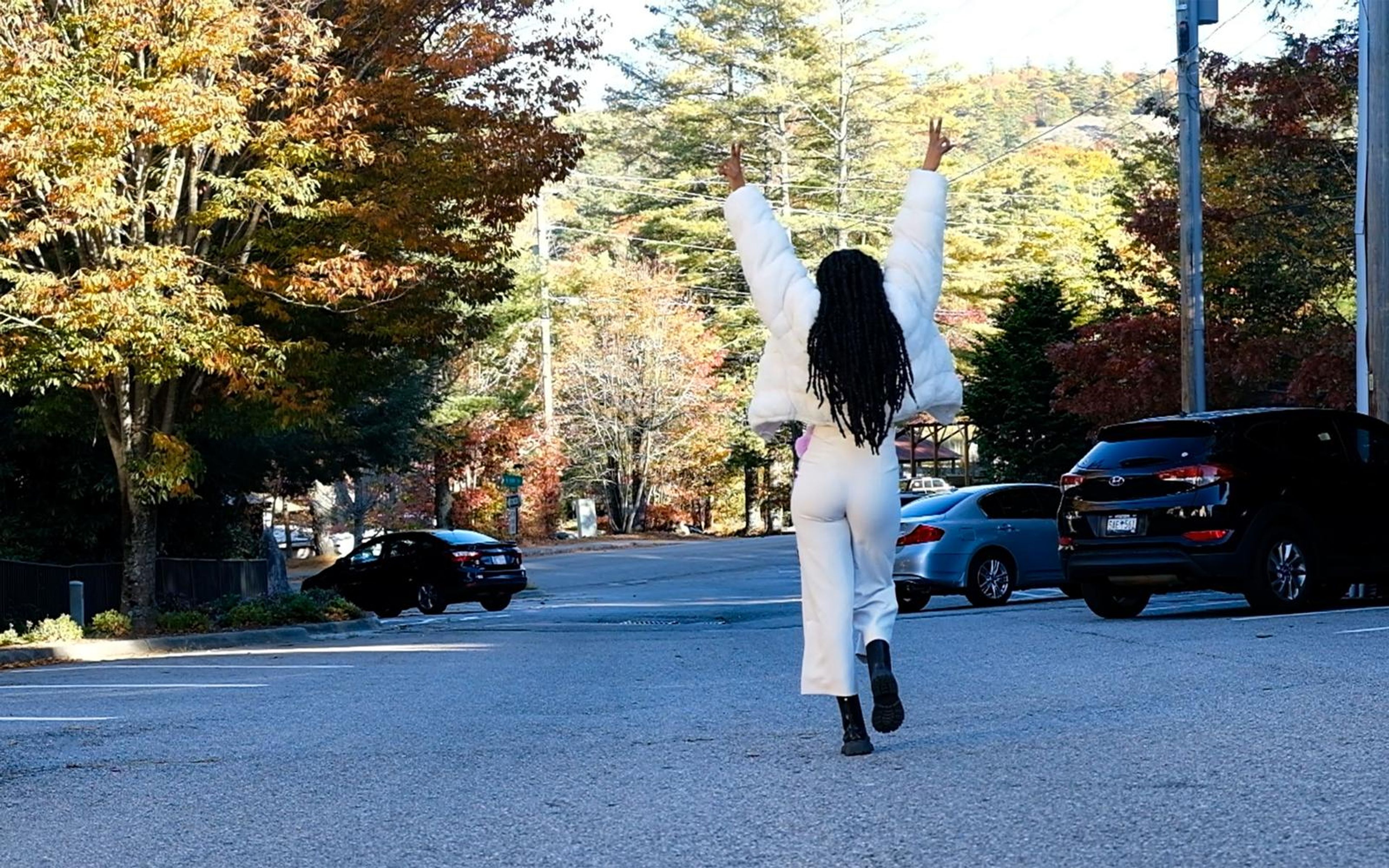 Model walking away from camera with both arms up in the street, white outfit, braids down her back