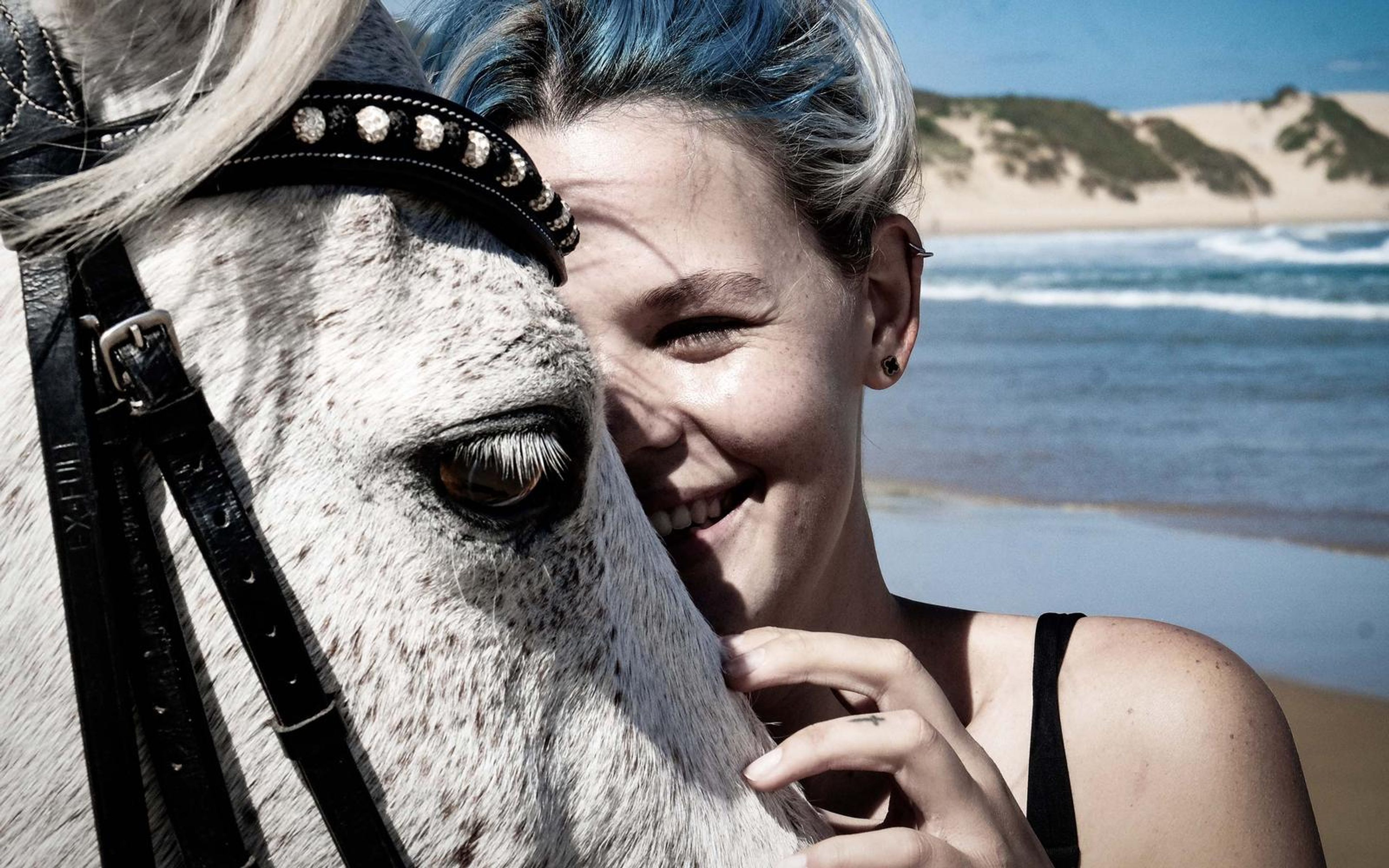 Woman smiling with her hand to her chin beside a bridled white horse, ocean in the background