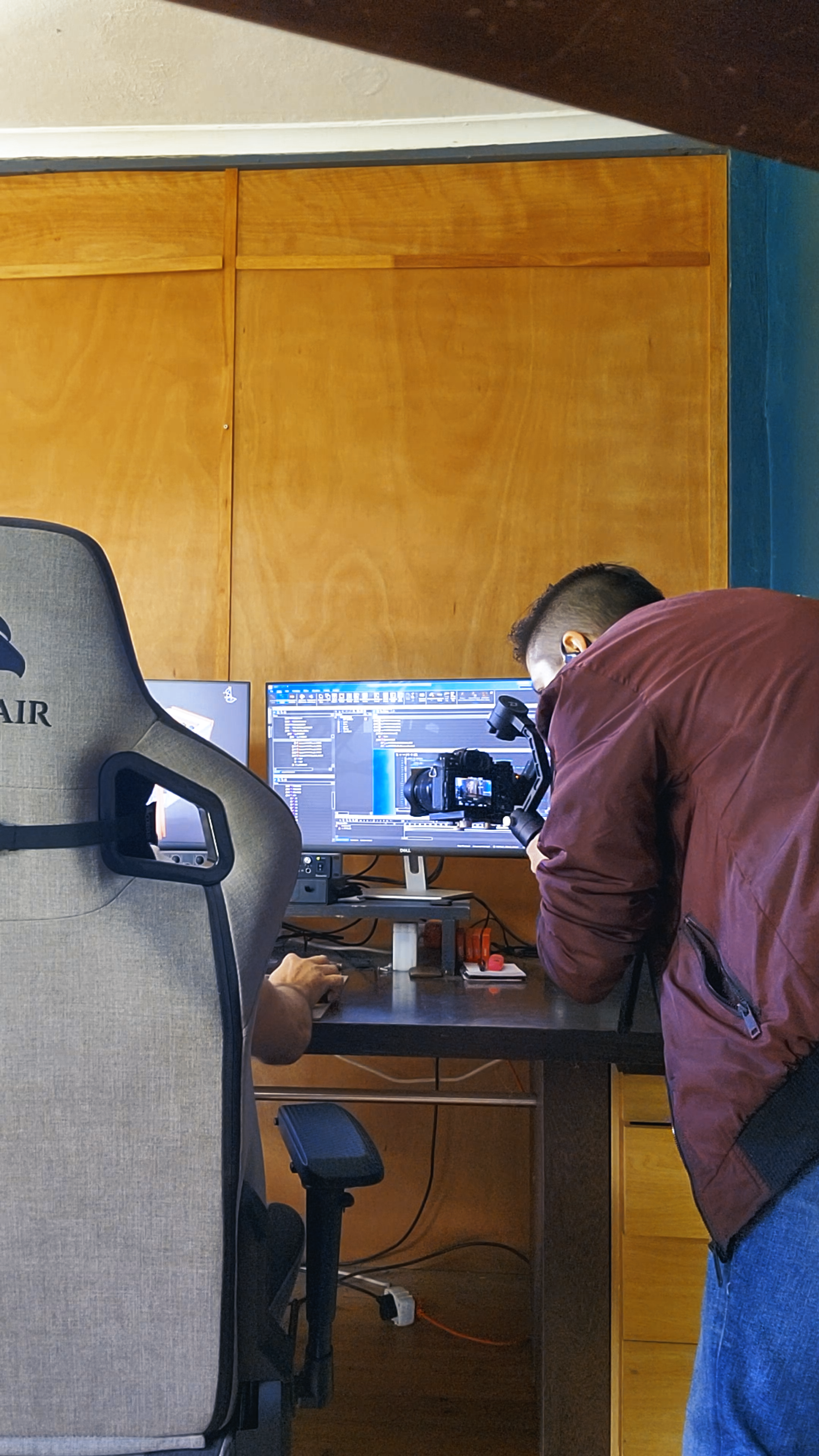 Behind the scenes, a man in a red jacket leaning over a desk at a monitor in a wood-panelled office