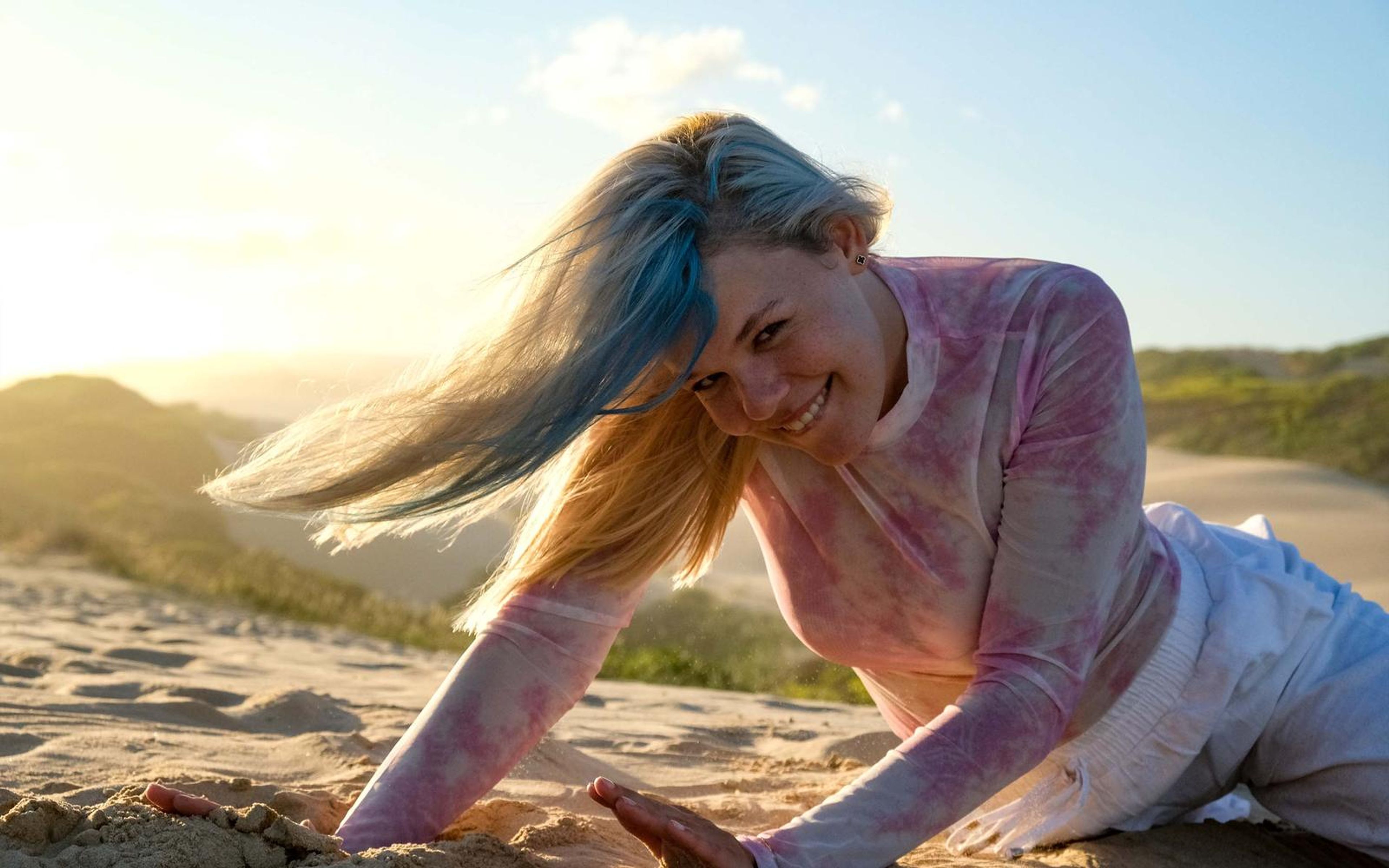 Woman smiling with her head tilted down and hair blowing, one hand in the sand