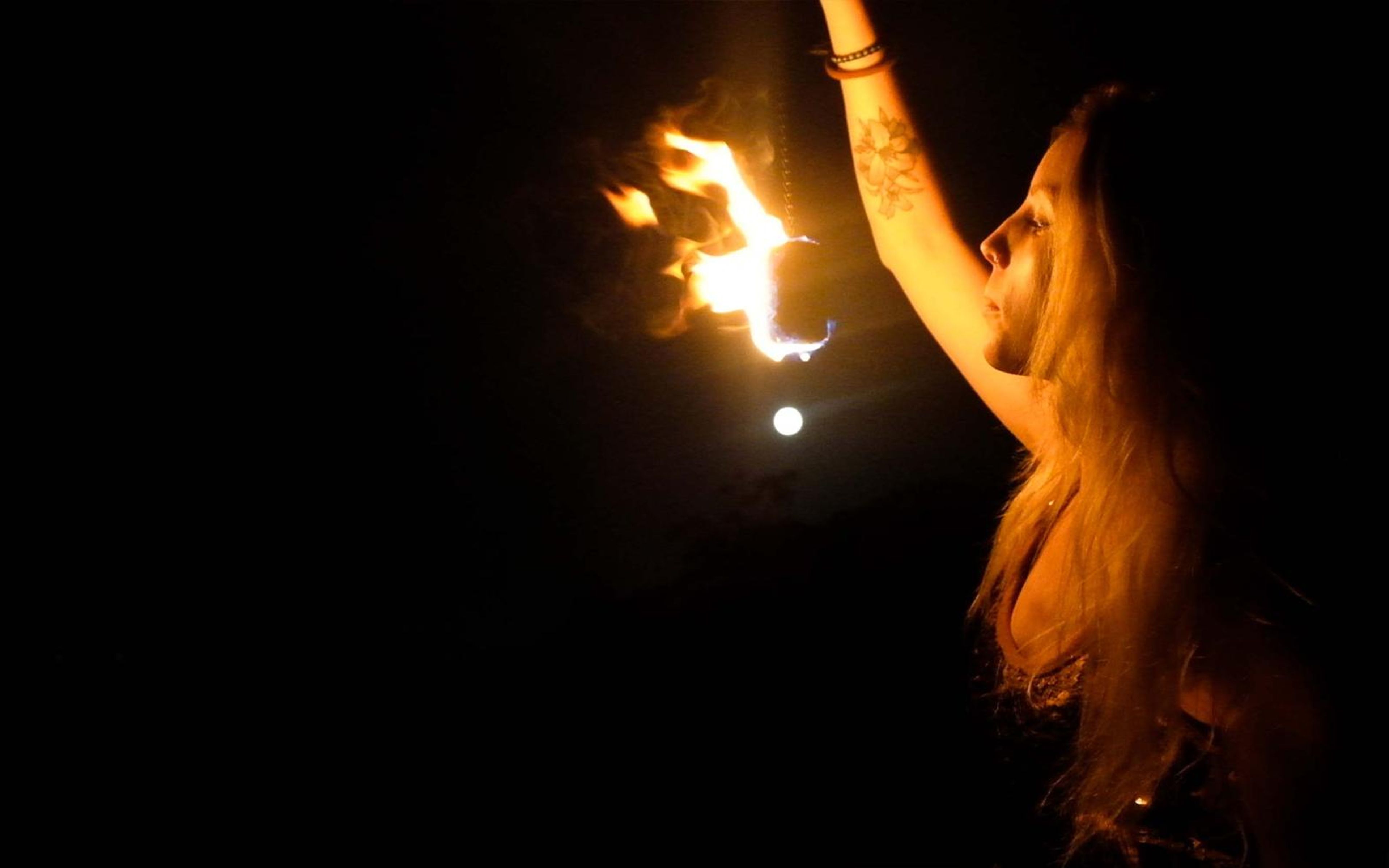 Woman looking upward with a fire prop blazing at upper left, gold hair lit from the side at night