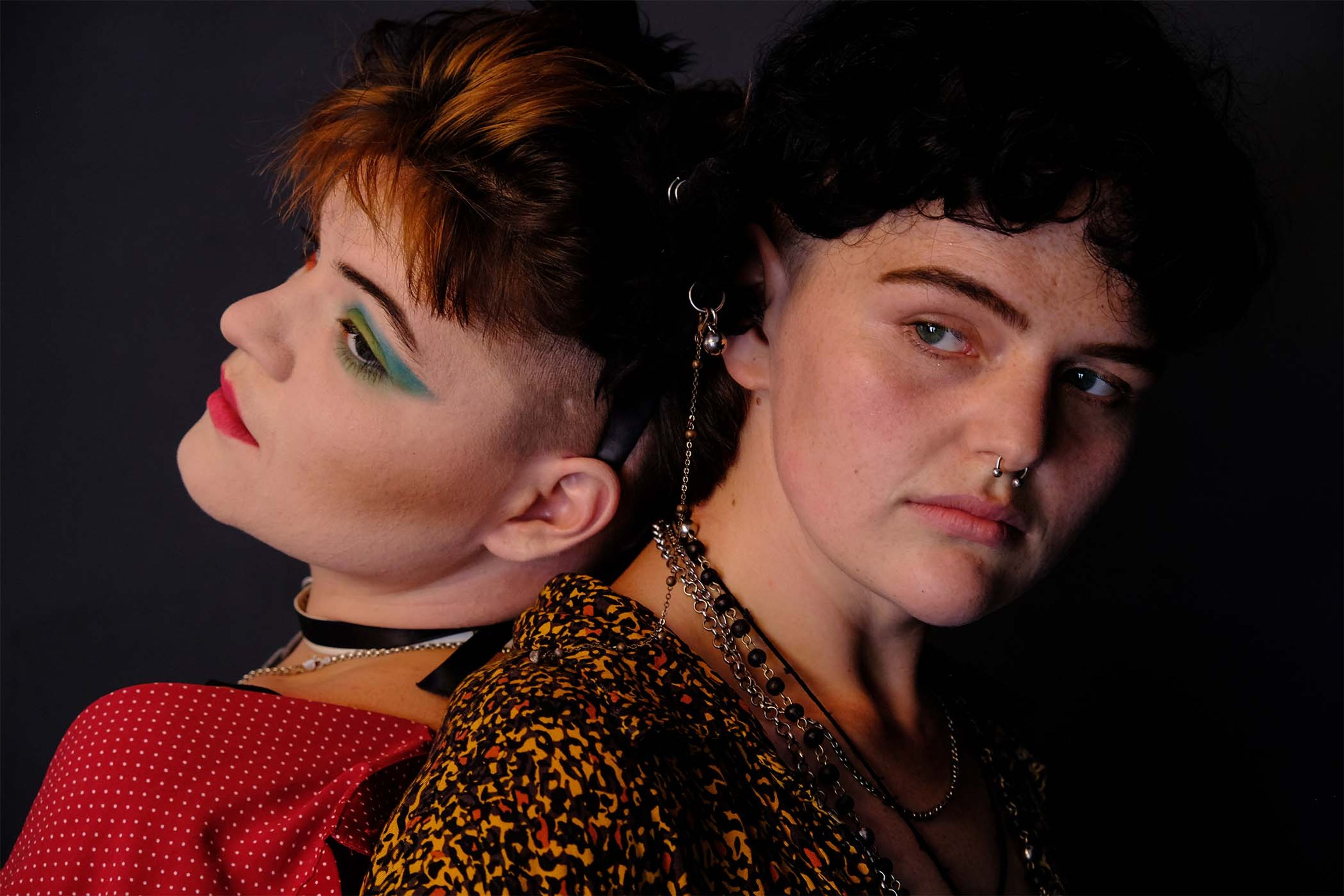 Back-to-back portrait of a couple, colorful eye makeup and patterned shirts, dark studio backdrop
