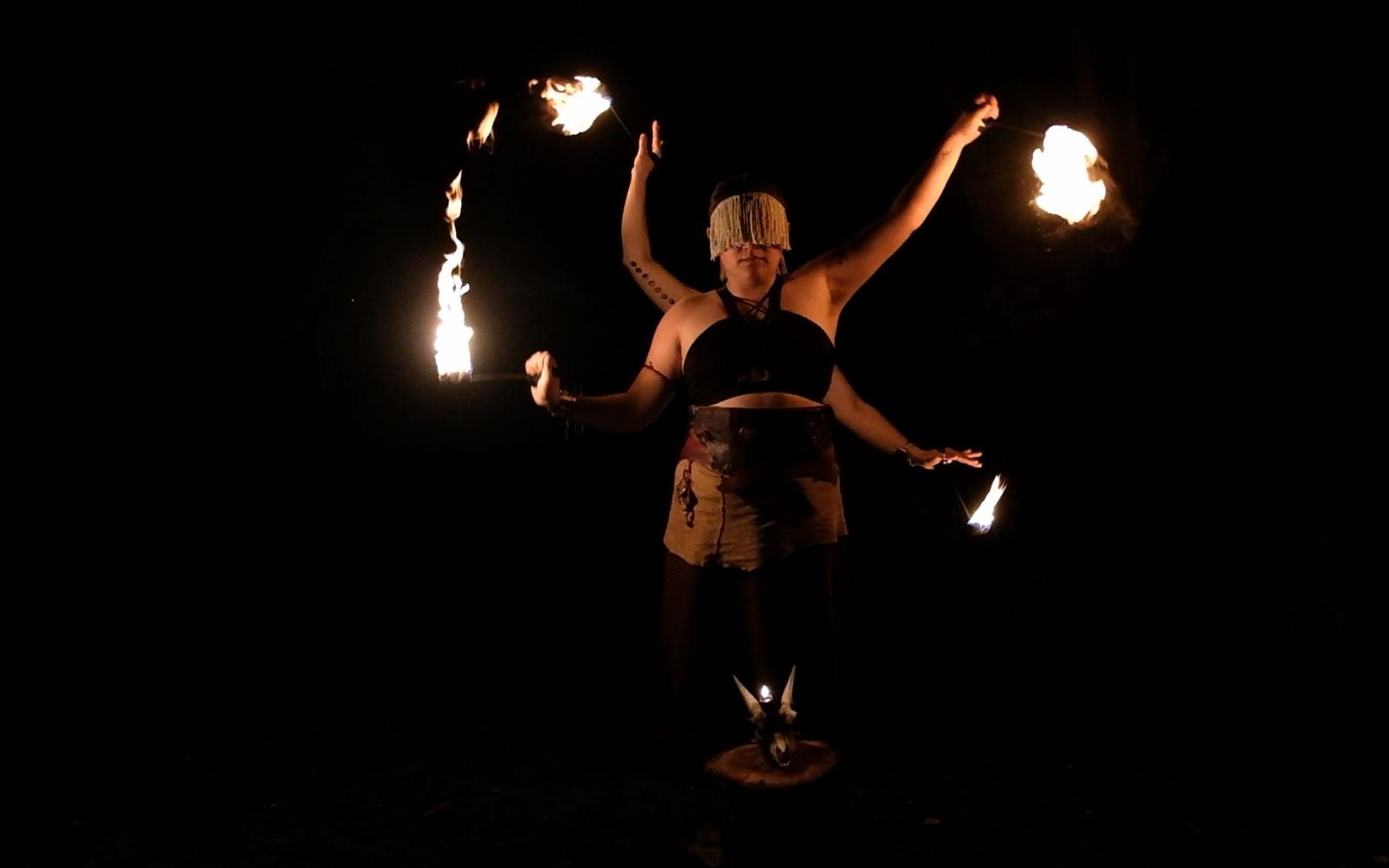 Performer in a fringed head covering spinning fire in arcs with arms spread, horned skull on the ground below
