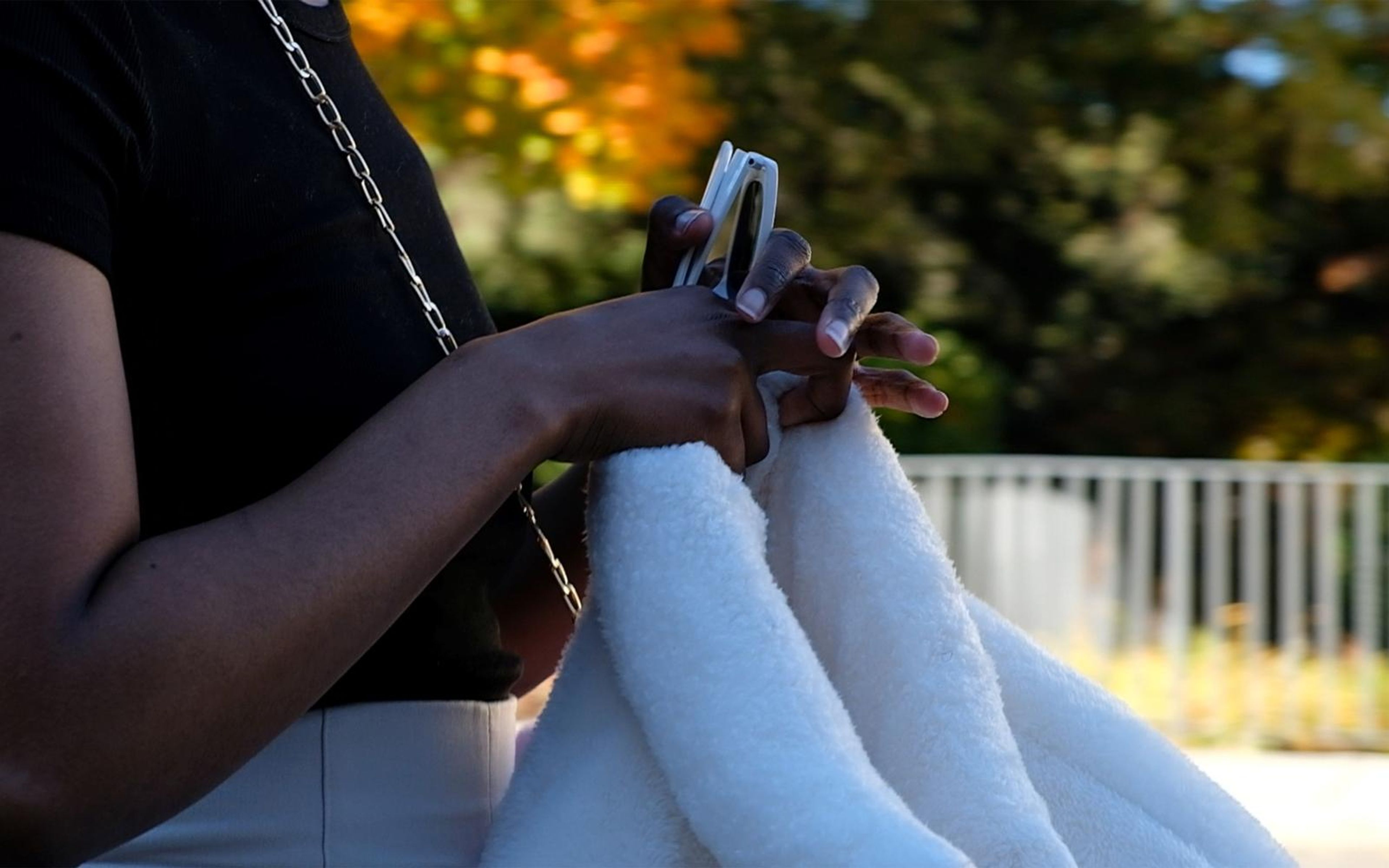 Close crop of hands holding white sunglasses beside a white fur jacket sleeve, black top with a chain strap