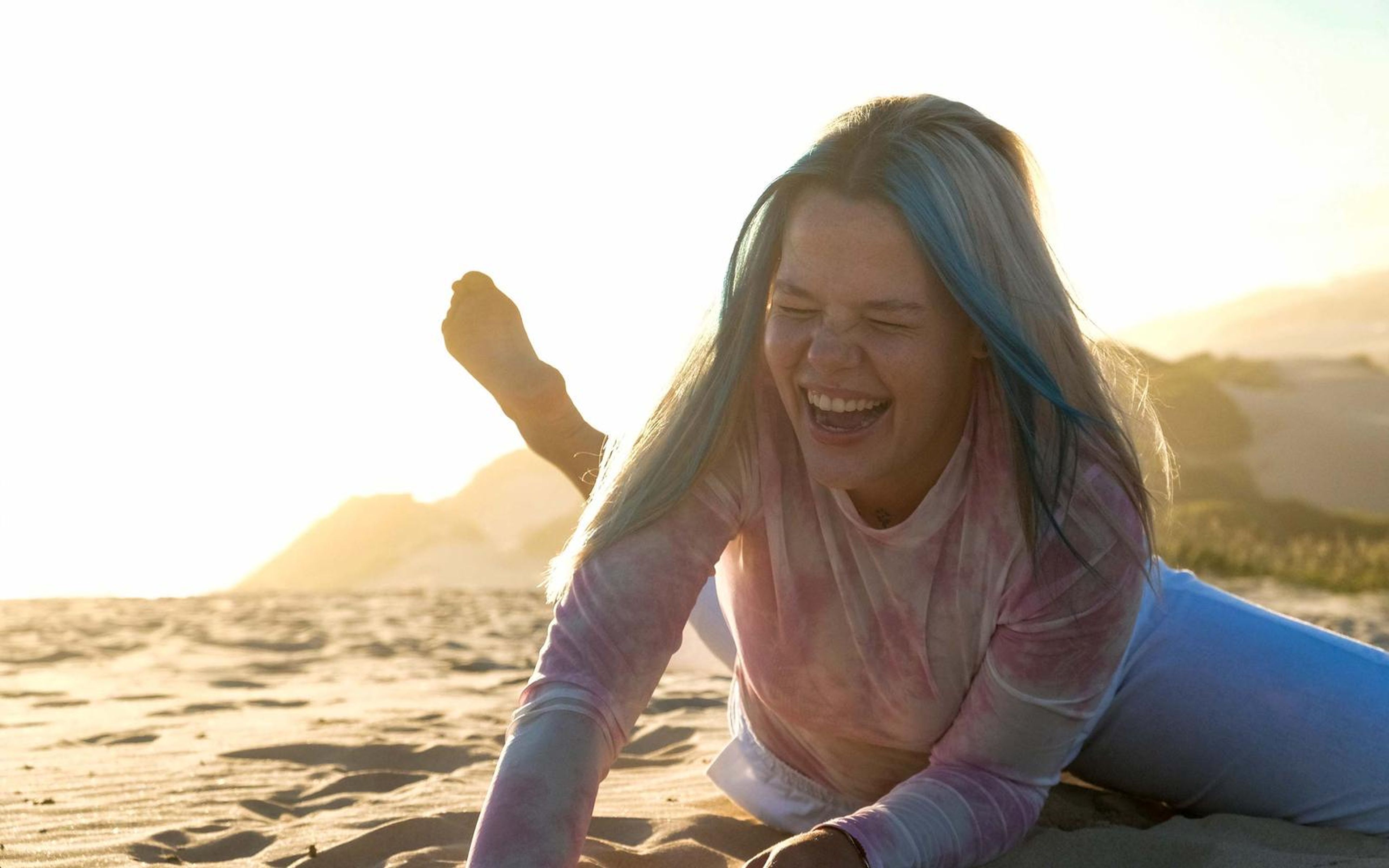 Woman laughing on the sand with one leg raised and blue-streaked hair across her face