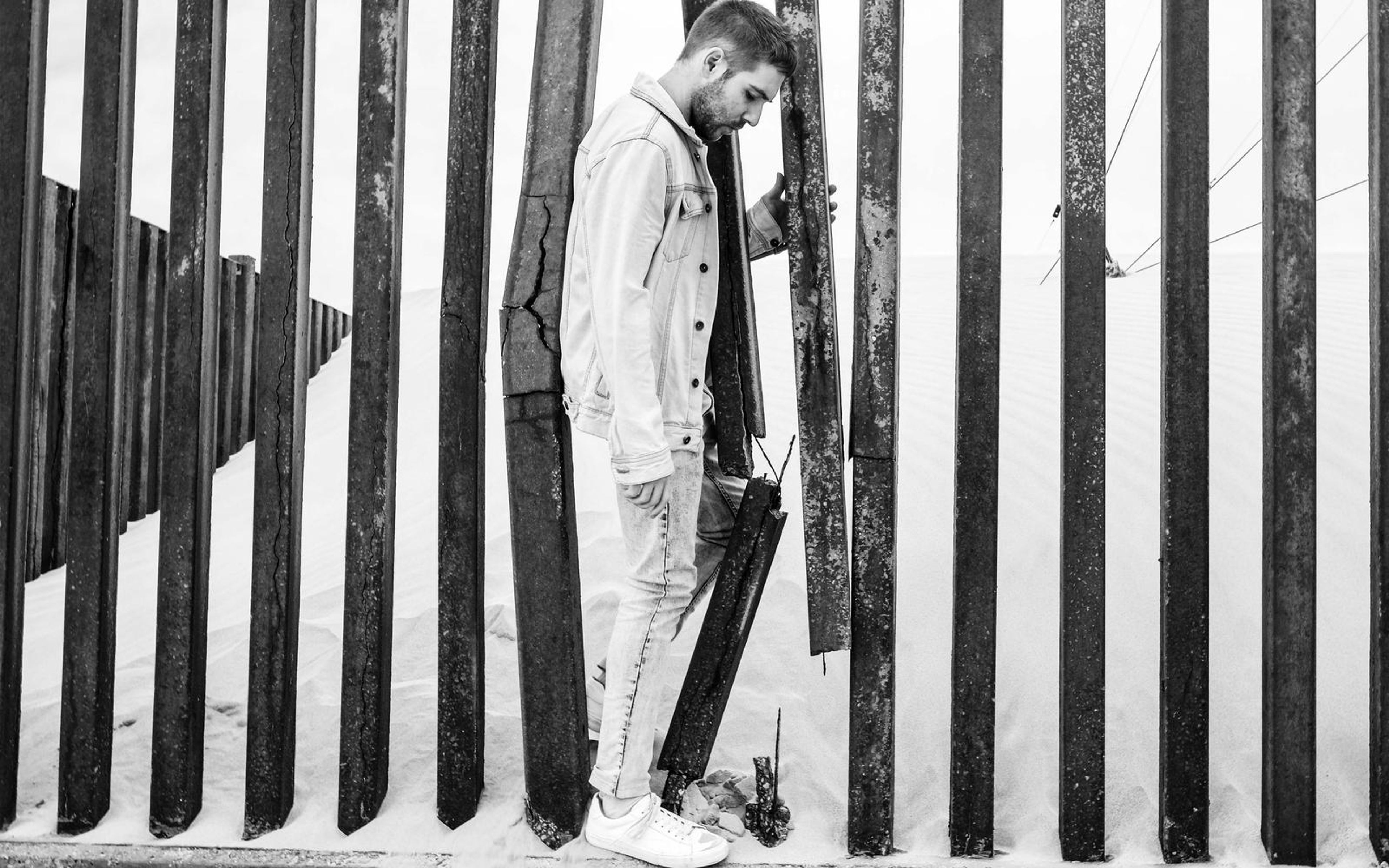 Black and white image of a man leaning on a gap in a wooden fence, sand dune behind, denim jacket