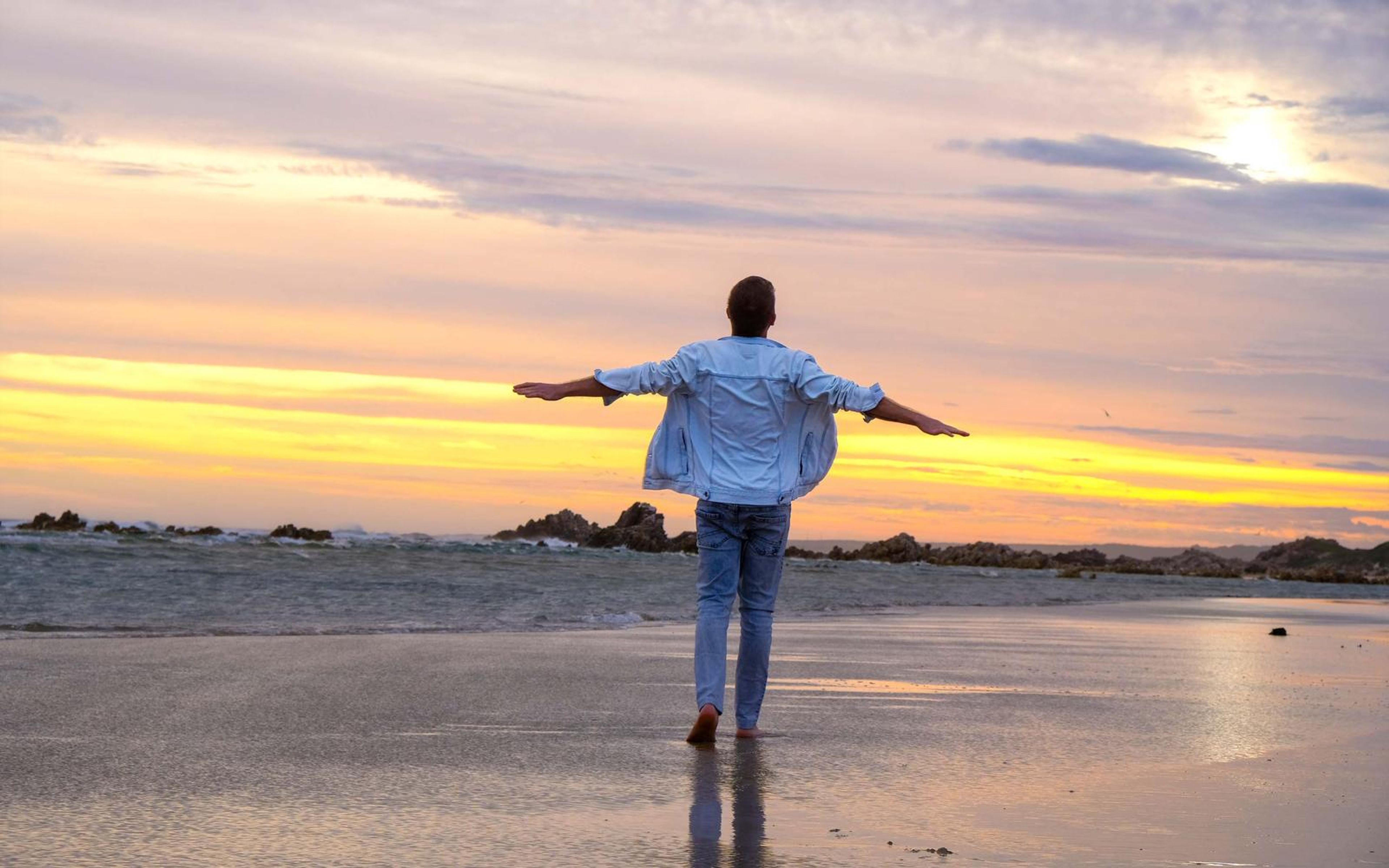 Man with arms outstretched facing a sunset sky over a wet beach, denim jacket open, barefoot