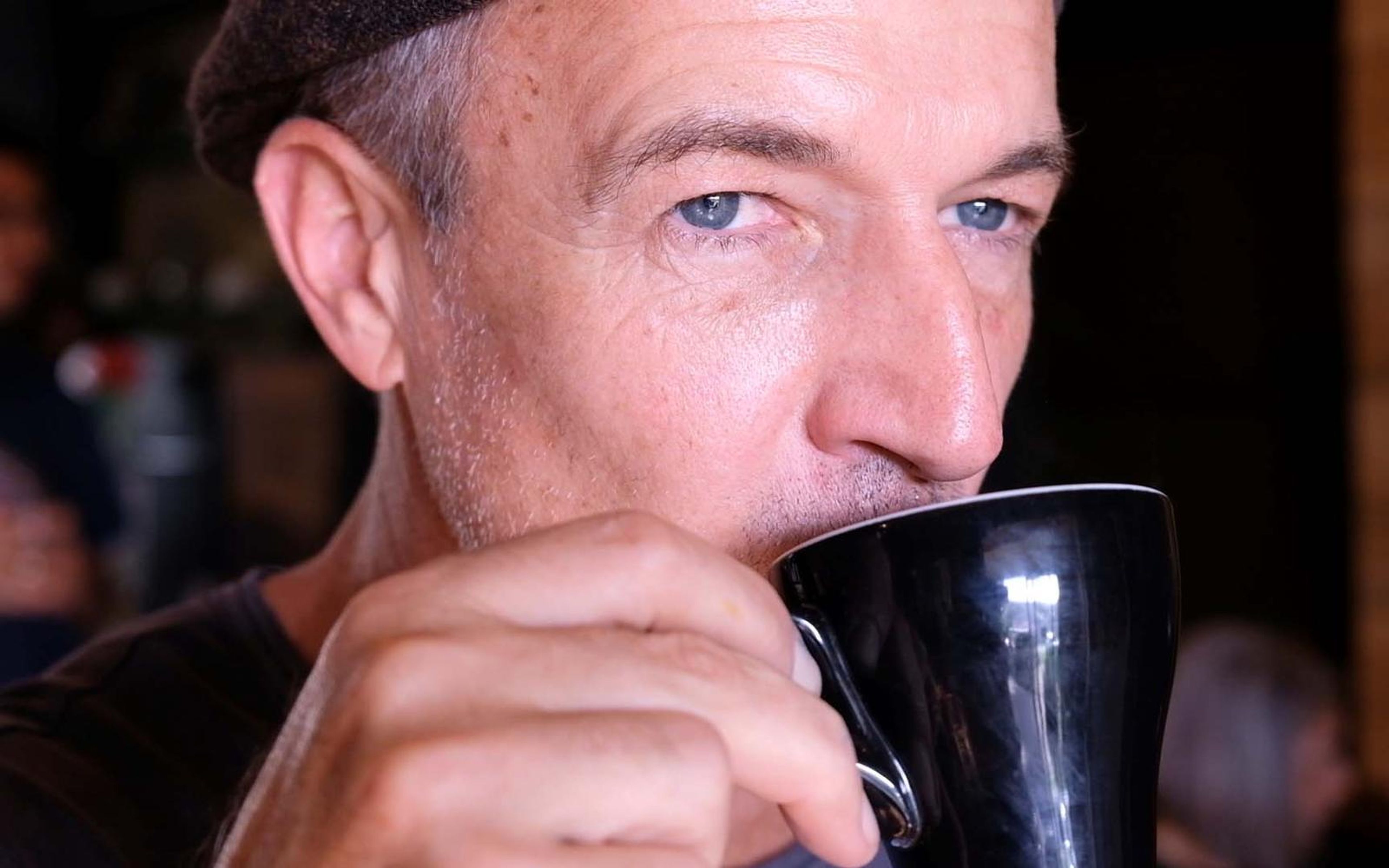Man in a flat cap with blue eyes drinking from a black mug with a cafe interior behind