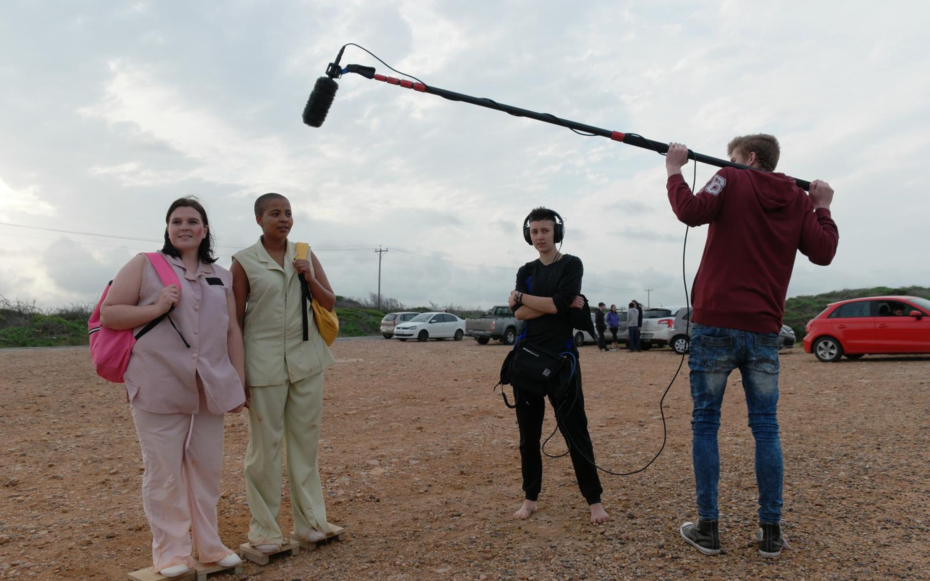 Two actors in pastel uniforms on a gravel lot with a boom operator and a crew member kneeling by a reflector