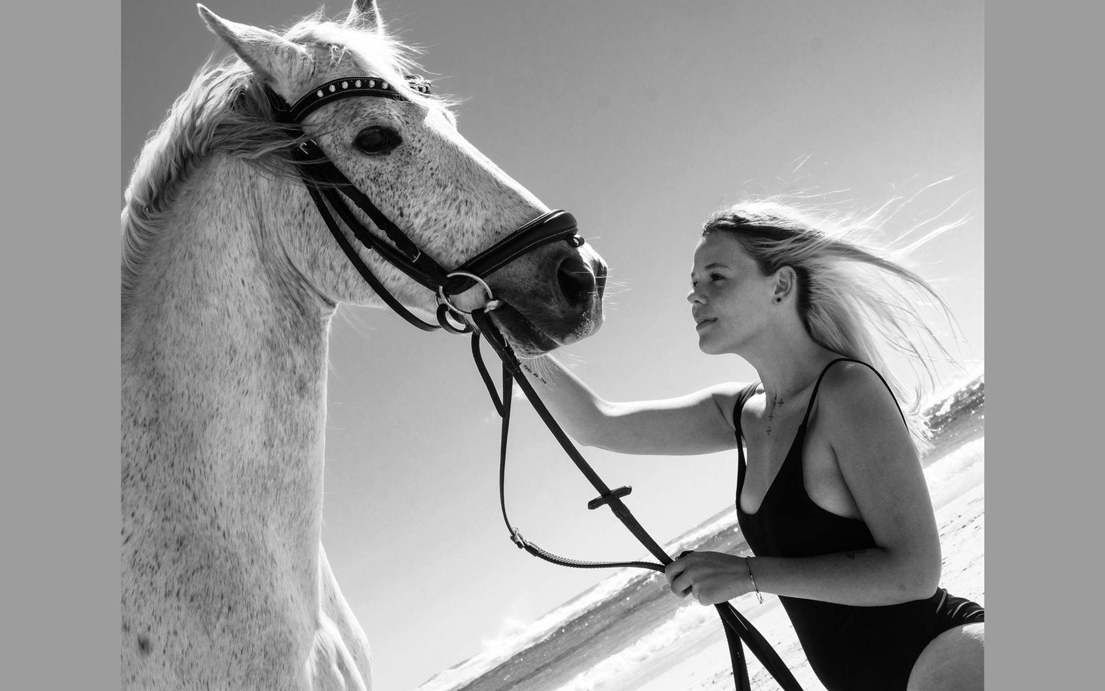 Black and white portrait of a woman reaching toward a horse's reins, bright sky behind