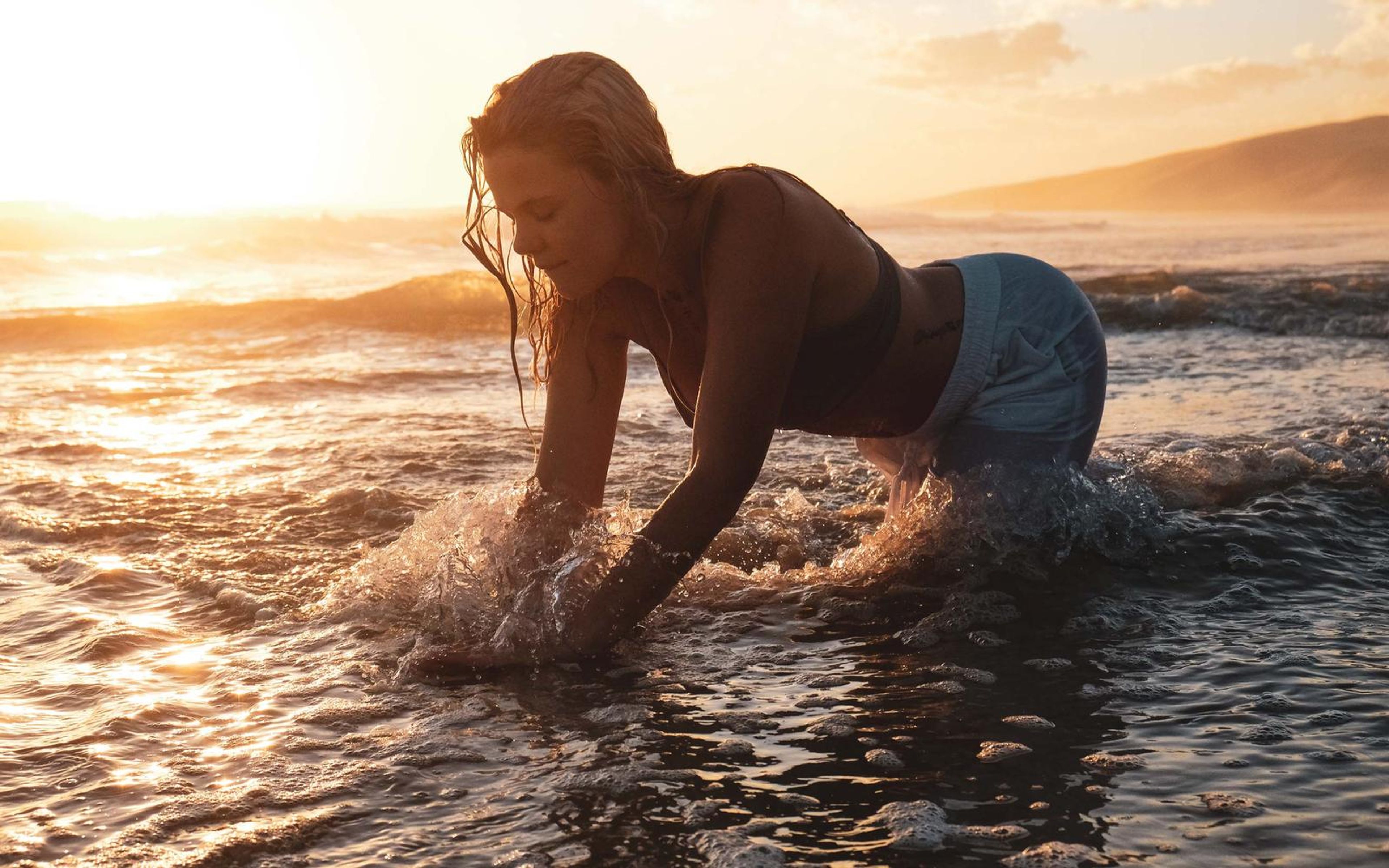 Woman crawling through the shallows with water splashing from her hands, wet hair, bright sunset behind